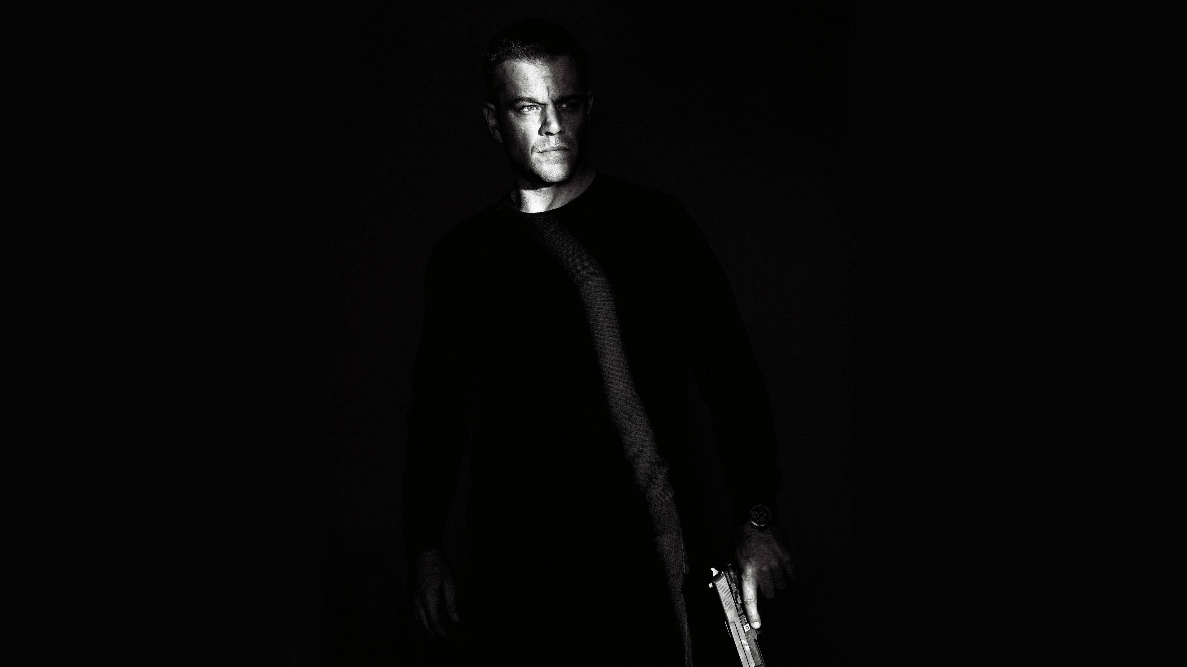 Matt Damon 桌布 壁紙 — 一個穿著深色襯衫的男人站在幾乎完全黑暗的環境中，手持一把槍，只有他的臉和手被微弱地照亮。