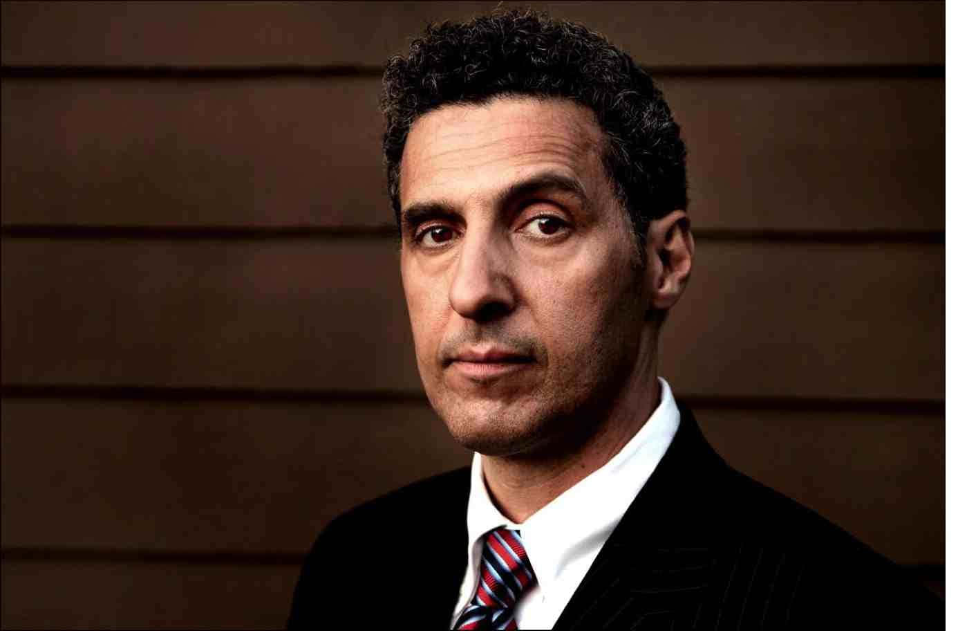 John Turturro 桌布 壁紙 — 一個穿著深色西裝和條紋領帶的嚴肅男人，在木鑲板背景前直視鏡頭。