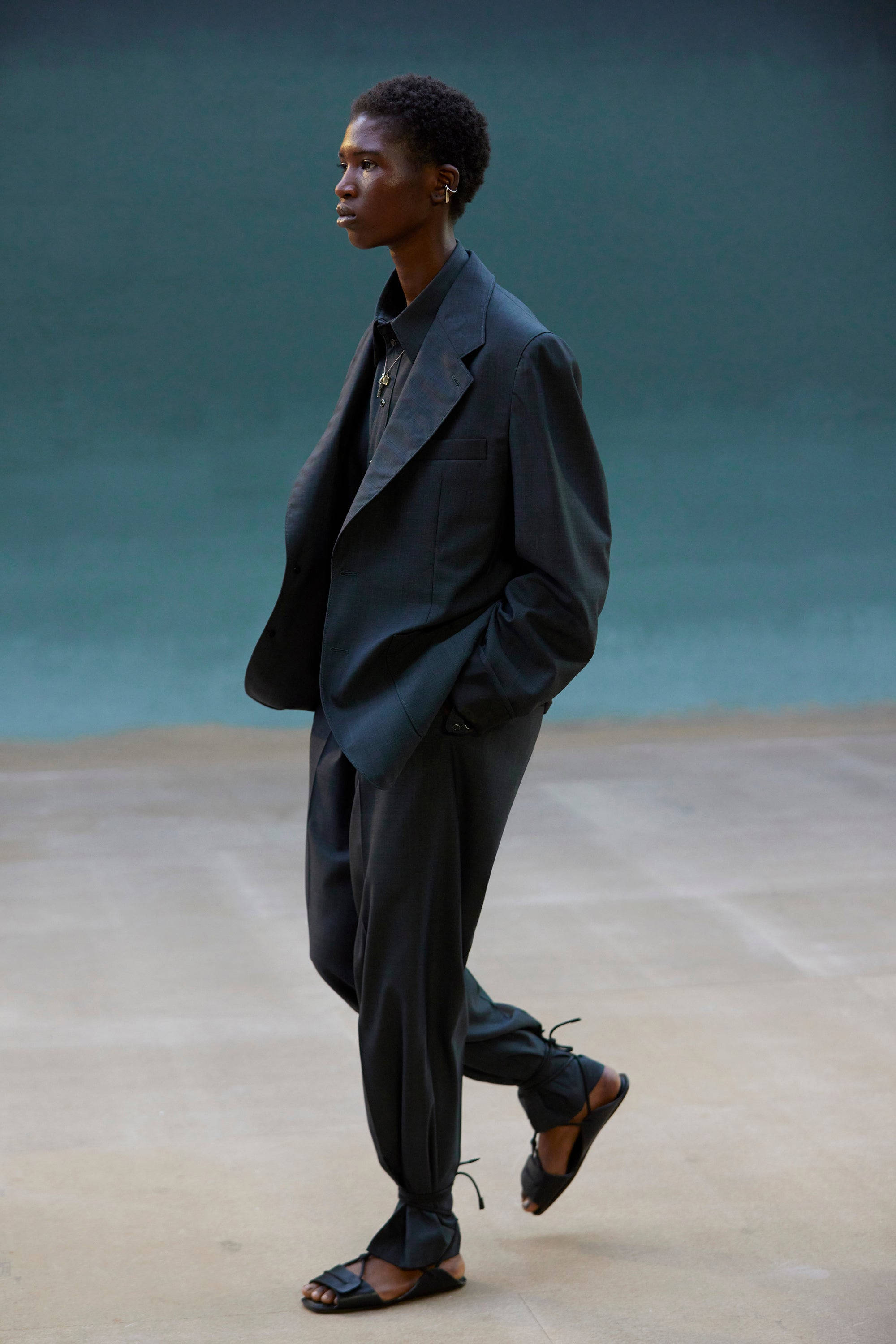 Mens Fashion 桌布 壁紙 — 一個穿著深色西裝和涼鞋的人，在藍綠色背景前自信地行走，具有極簡美學。