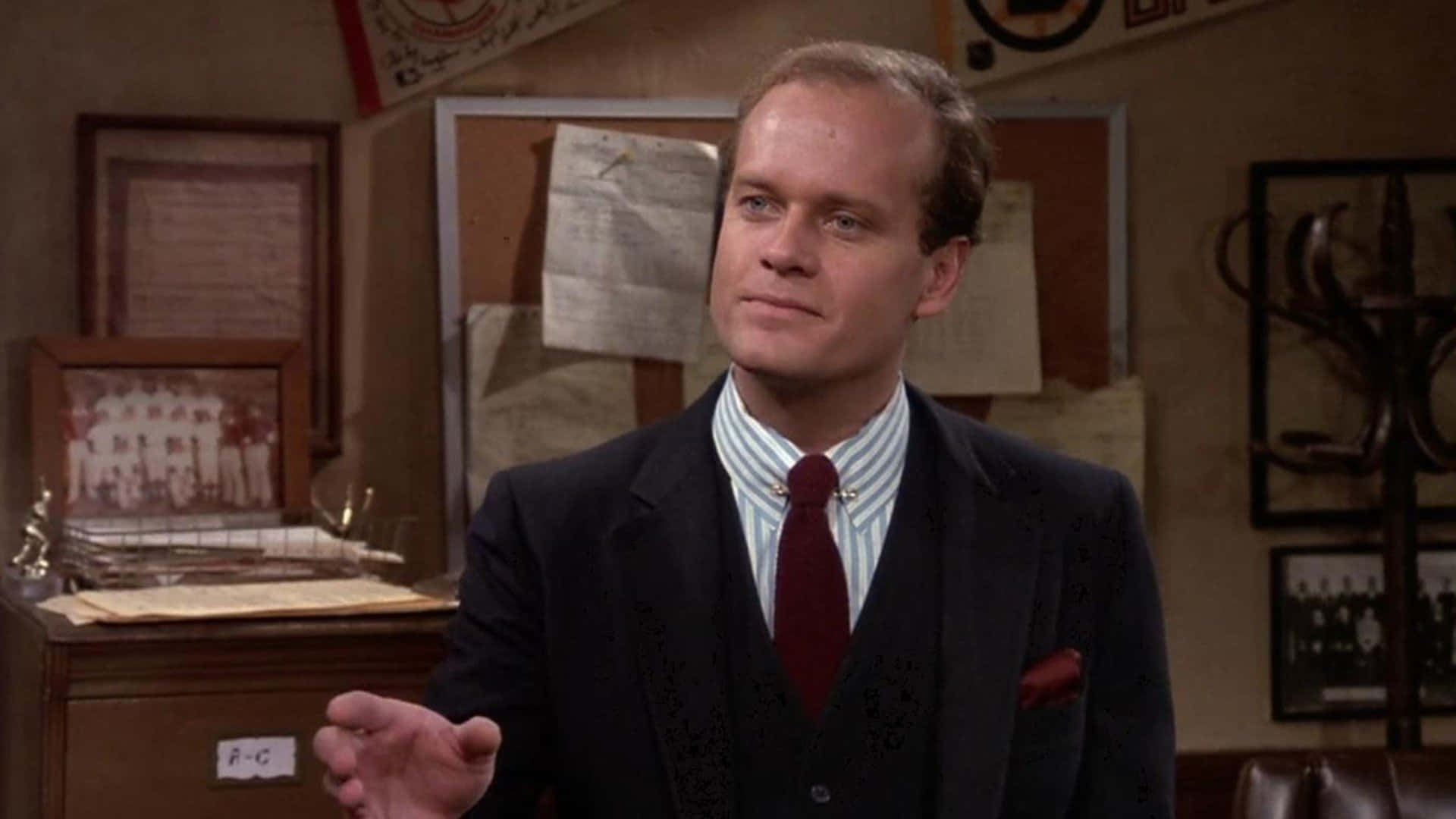 Kelsey Grammer 桌布 壁紙 — 一個穿著深色西裝和紅色領帶的男人站在一個堆滿相框照片和紀念品的凌亂辦公室裡。