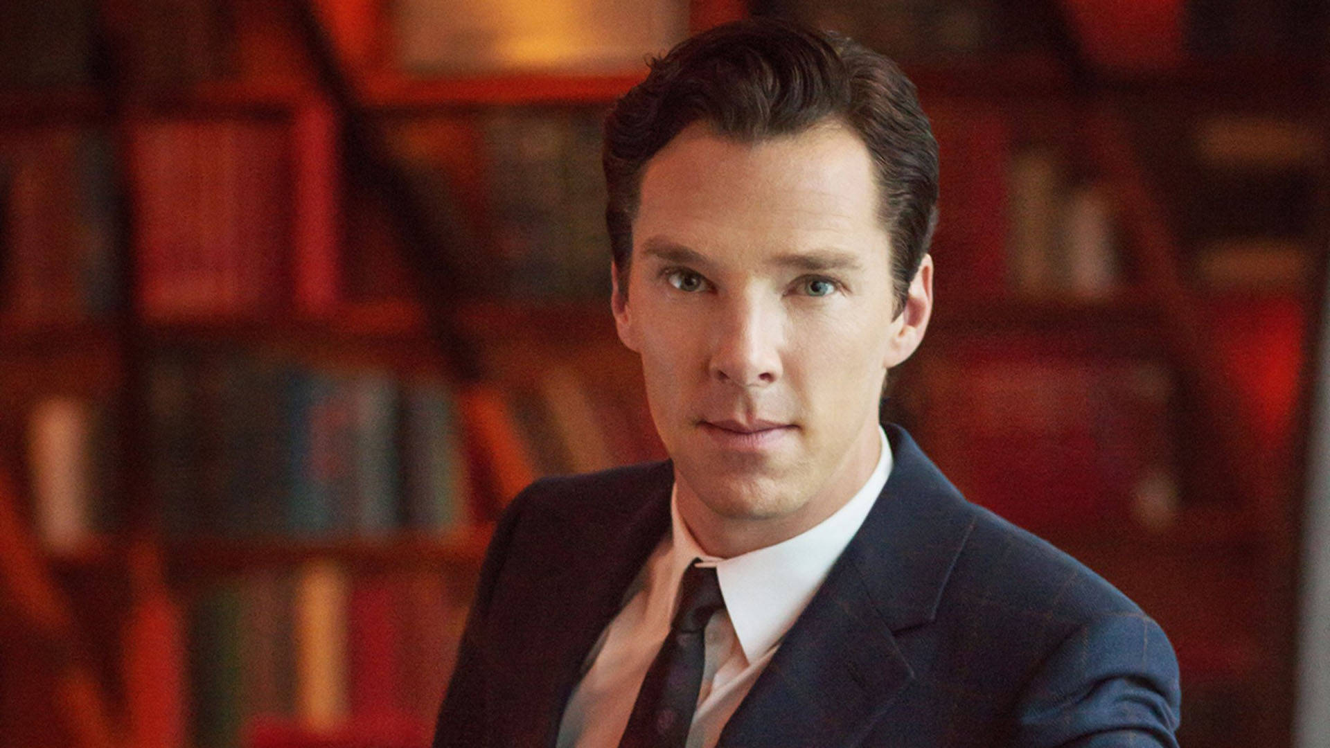 benedict 桌布 壁紙 — 一個穿著深色西裝和領帶的男人，在溫暖照明的圖書館中凝視，身後是模糊的書架。