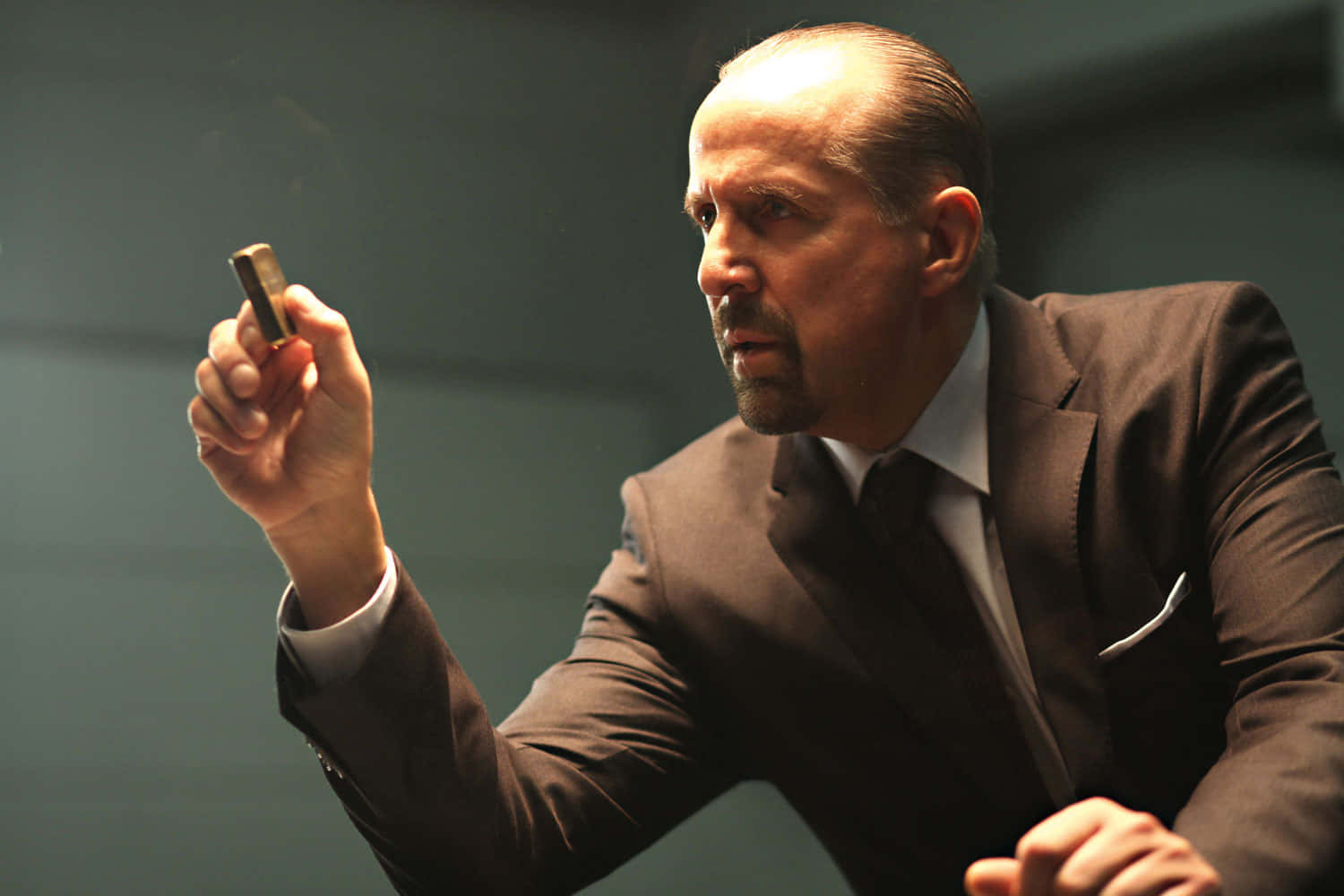 Peter Stormare 桌布 壁紙 — 一個穿著深色西裝的男人，在柔和的灰色背景下，專注地拿著一個小物件。