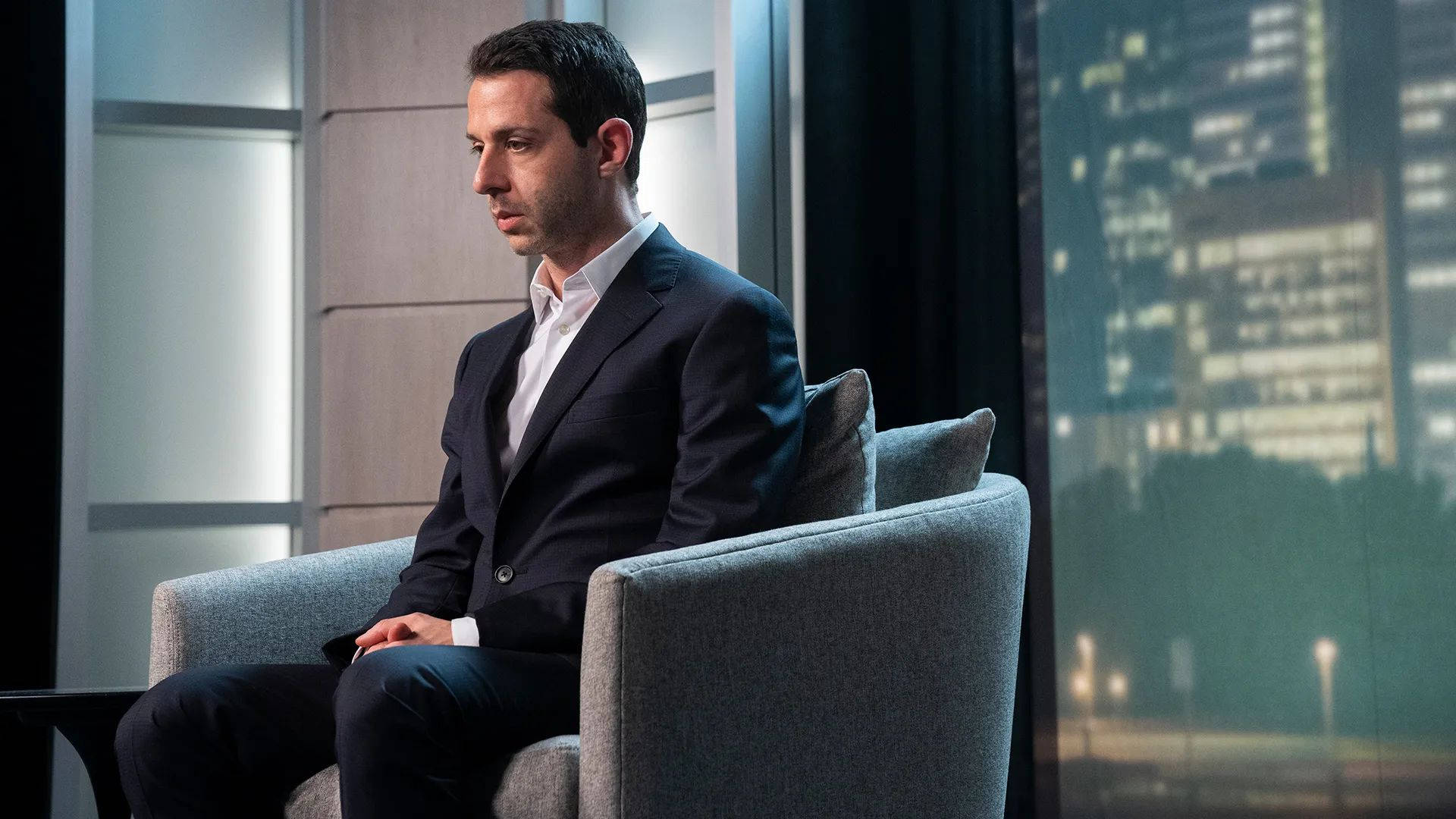 Succession 桌布 壁紙 — 一個穿著深色西裝的男人在現代扶手椅上沉思，背景是夜晚的城市景觀。