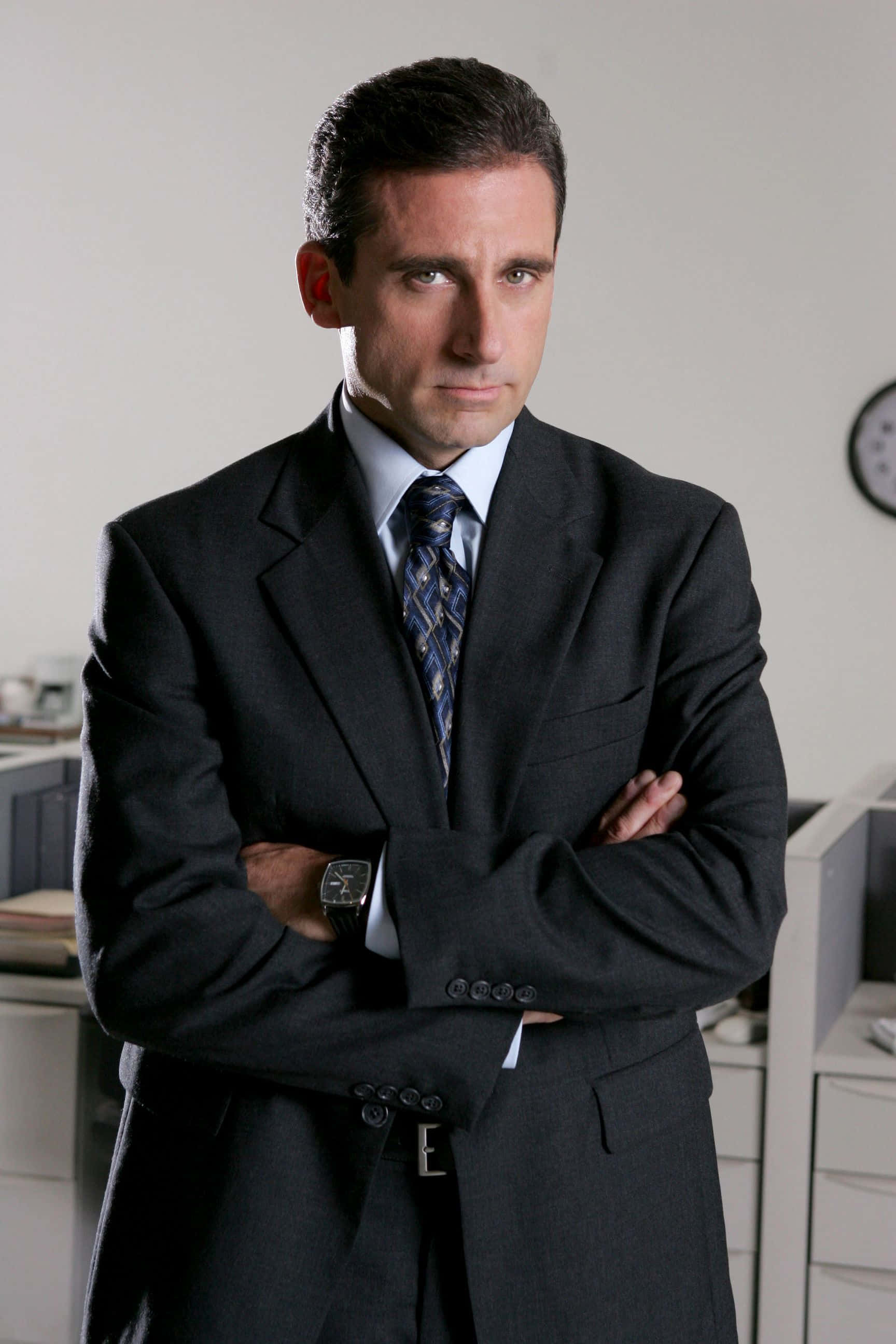 Michael Scott 桌布 壁紙 — 一個穿著深色西裝的男人在辦公室交叉雙臂站立，嚴肅地看著鏡頭。