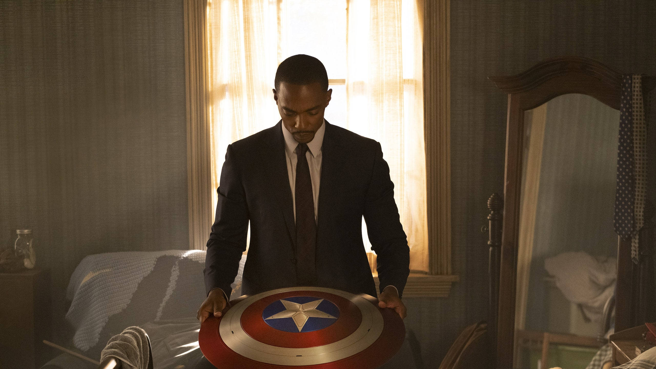 Anthony Mackie 桌布 壁紙 — 一個穿著深色西裝的男人在陽光普照的復古臥室中手持美國隊長的盾牌，房間內有條紋壁紙和木製家具。