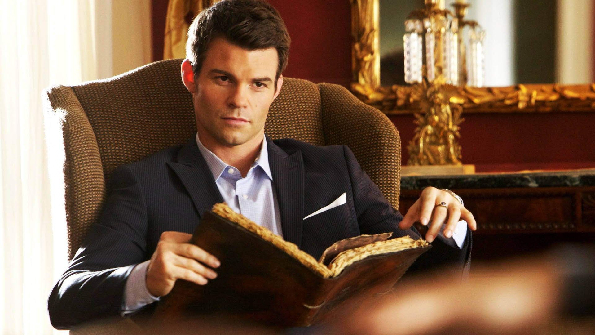elijah mikaelson 桌布 壁紙 — 一個穿著深色西裝的男人坐在一張豪華扶手椅上，在一個裝飾華麗、帶有金色點綴的房間裡拿著一本舊書。
