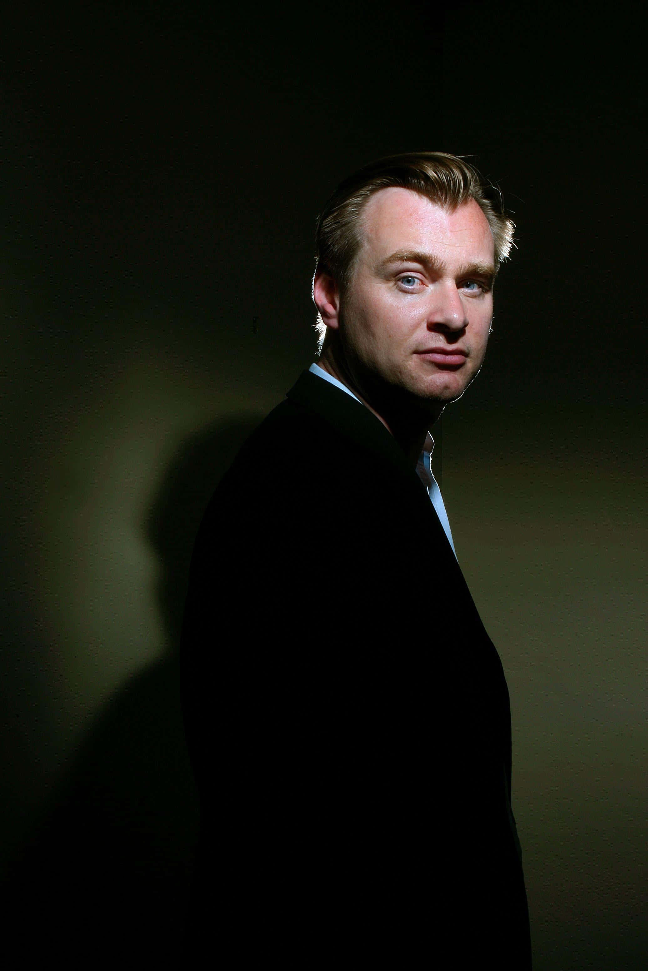 Christopher Nolan 桌布 壁紙 — 一個穿著深色西裝的男人站在陰影的攝影棚燈光下，神情嚴肅，帶著一絲微笑。