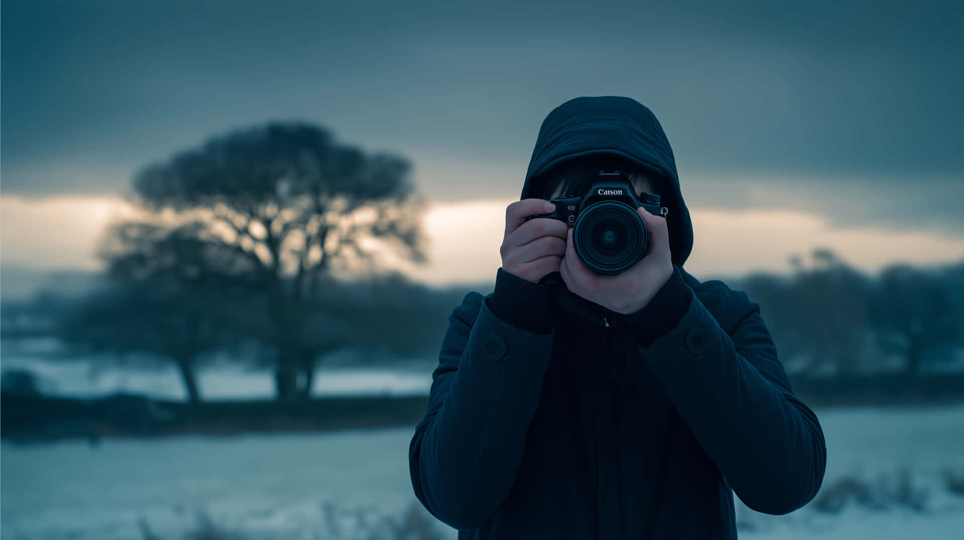 Mysterious Photographer in Hoody with Camera Winter Setting 壁紙 — 一個穿著深色連帽外套的人，在雪地上舉起相機對著臉，背景是光禿禿的樹木和陰沉的暮色天空。