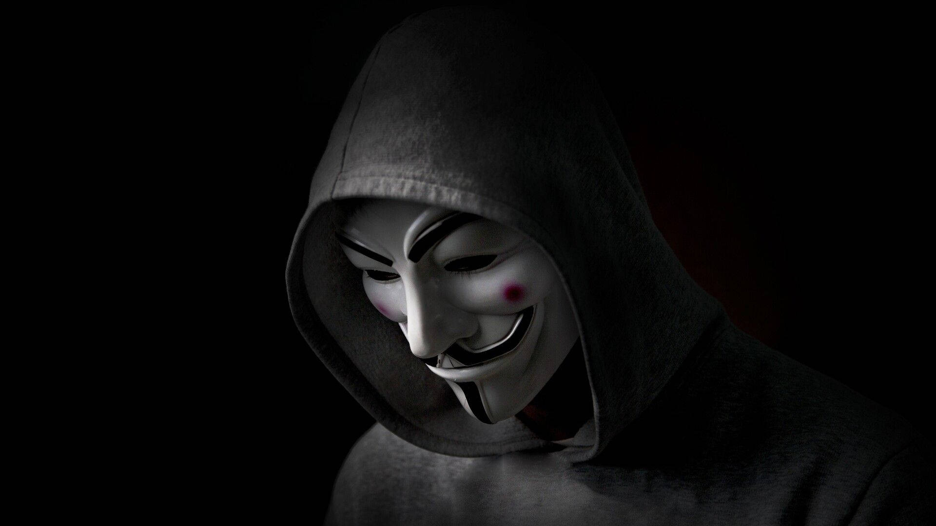 Hacker Full HD 桌布 壁紙 — 一個穿著深色連帽衫的人，在黑色背景前戴著白色蓋伊·福克斯面具，臉頰紅色。