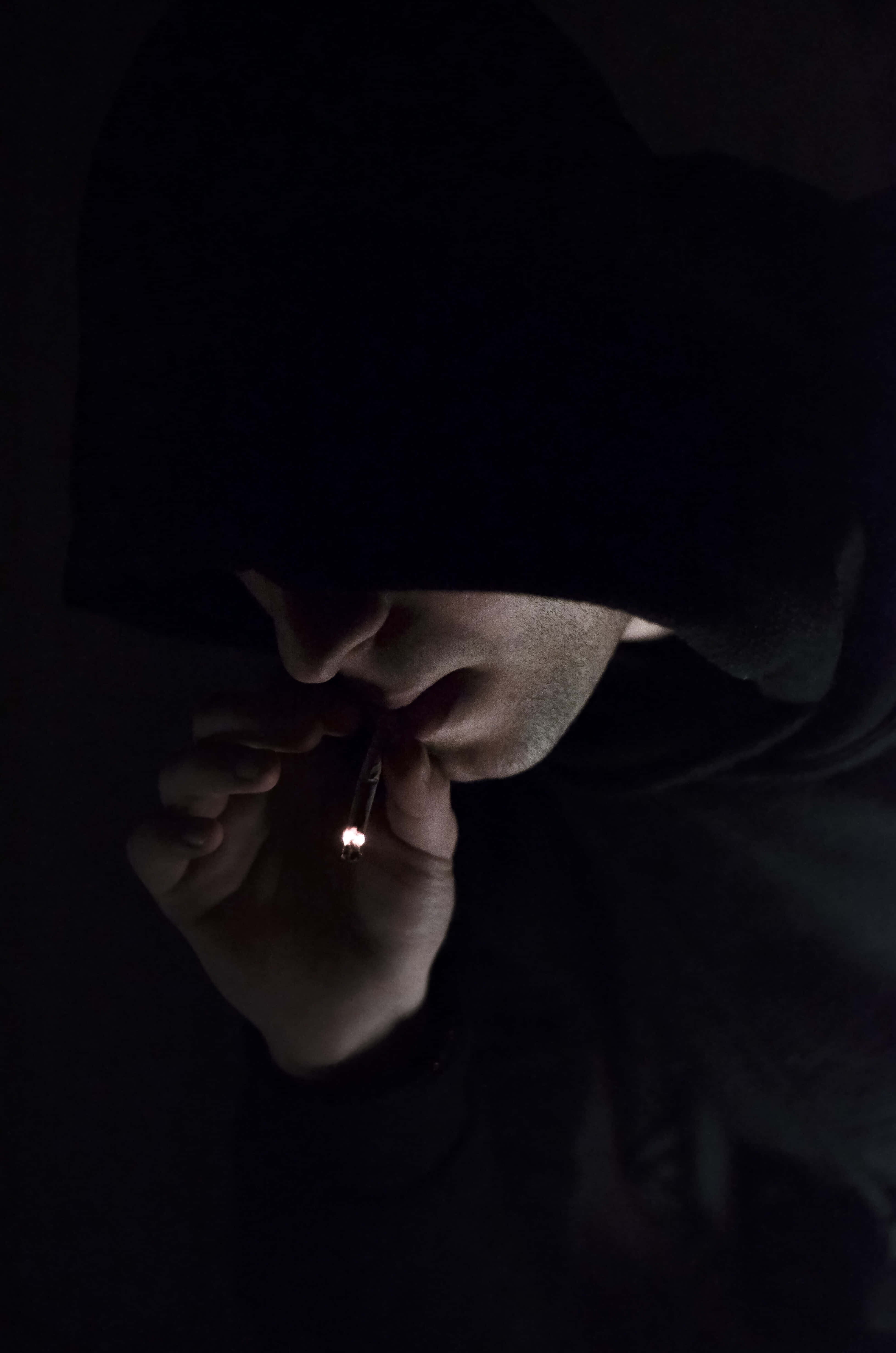 桌布 壁紙 — 一個穿著深色連帽衫的人在幾乎完全黑暗的環境中點燃一支香煙，只有火焰在他們的臉上投下戲劇性的陰影。