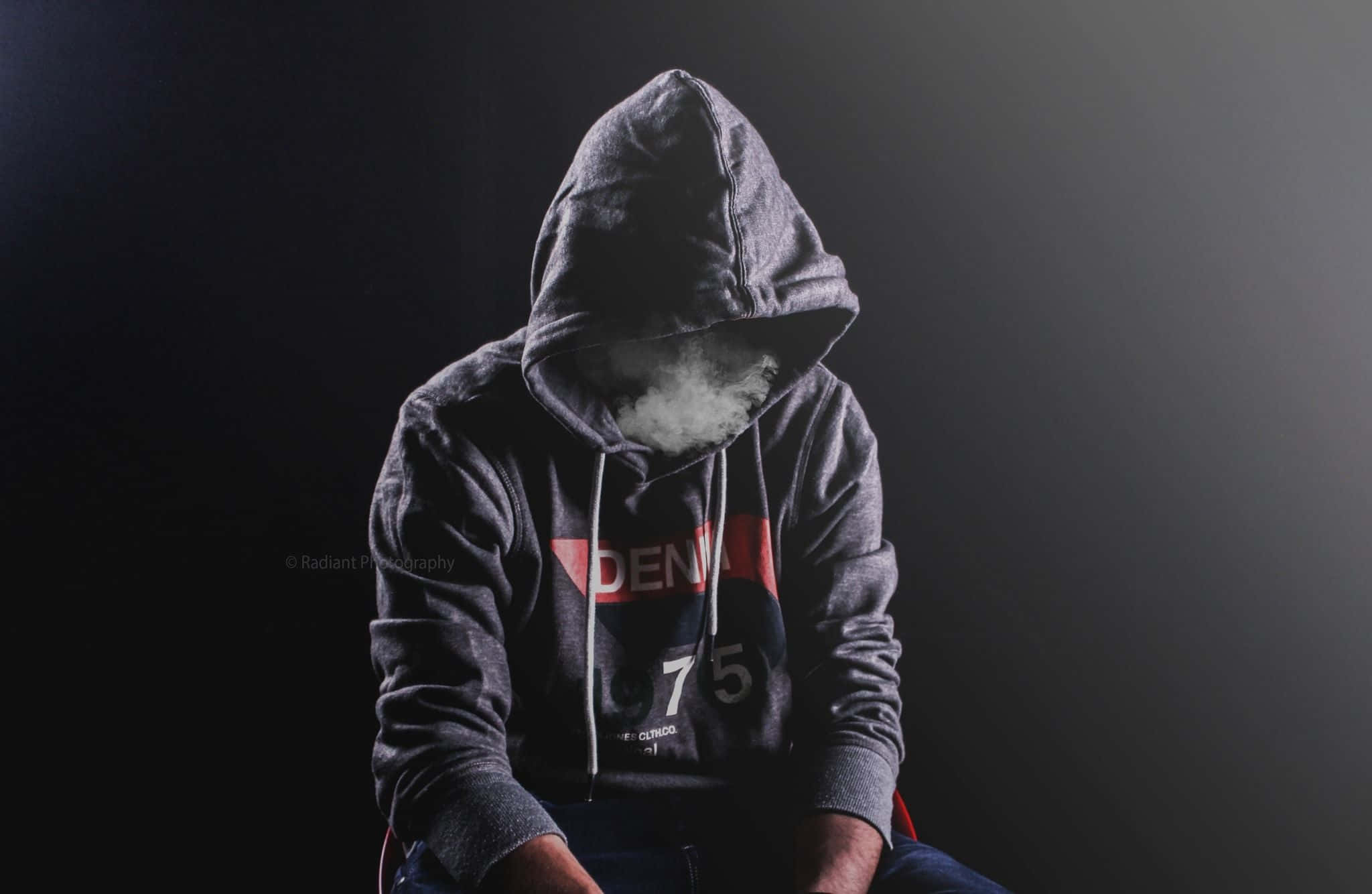 Hoodie 桌布 壁紙 — 一個穿著深色連帽衫的人彎腰坐著，煙霧遮住了他們的臉，背景是黑色。