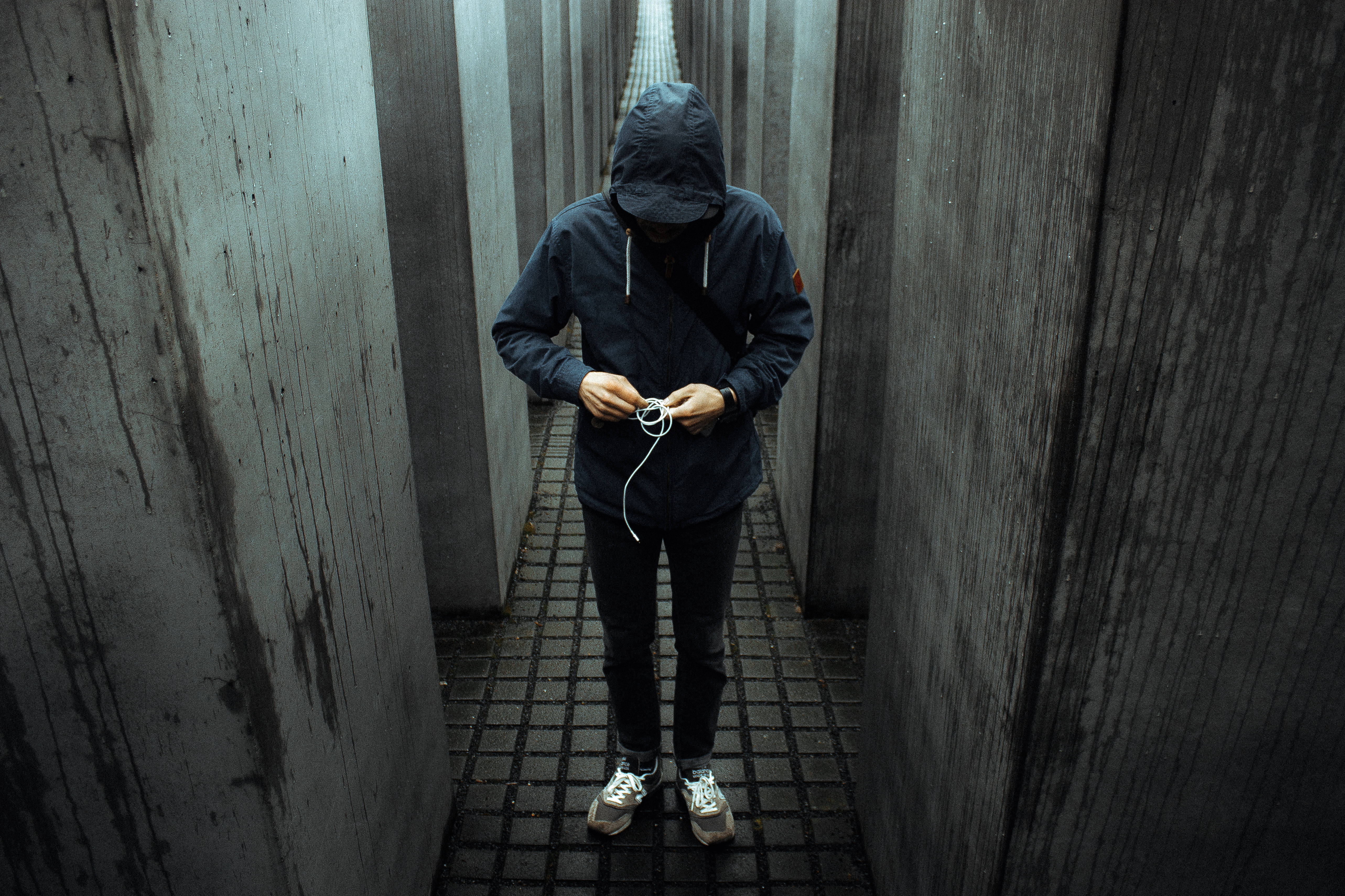 Forever Alone 桌布 壁紙 — 一個穿著深色連帽衫的人獨自站在狹窄的混凝土走廊裡，擺弄著耳塞。
