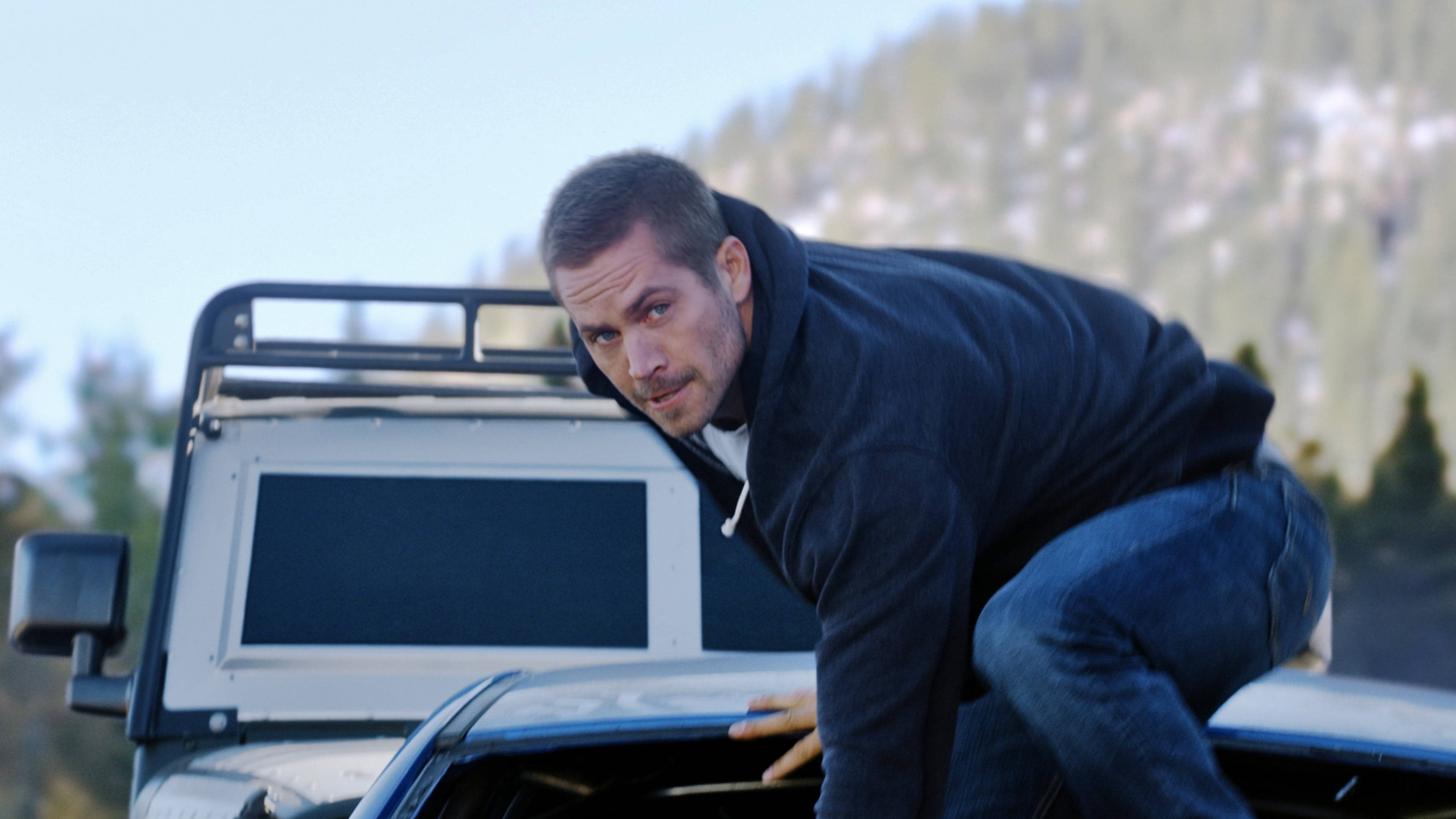 Paul Walker 桌布 壁紙 — 一個穿著深色連帽衫的男人靠在一輛堅固卡車的引擎蓋上，背景是晴朗天空下白雪覆蓋的山脈。
