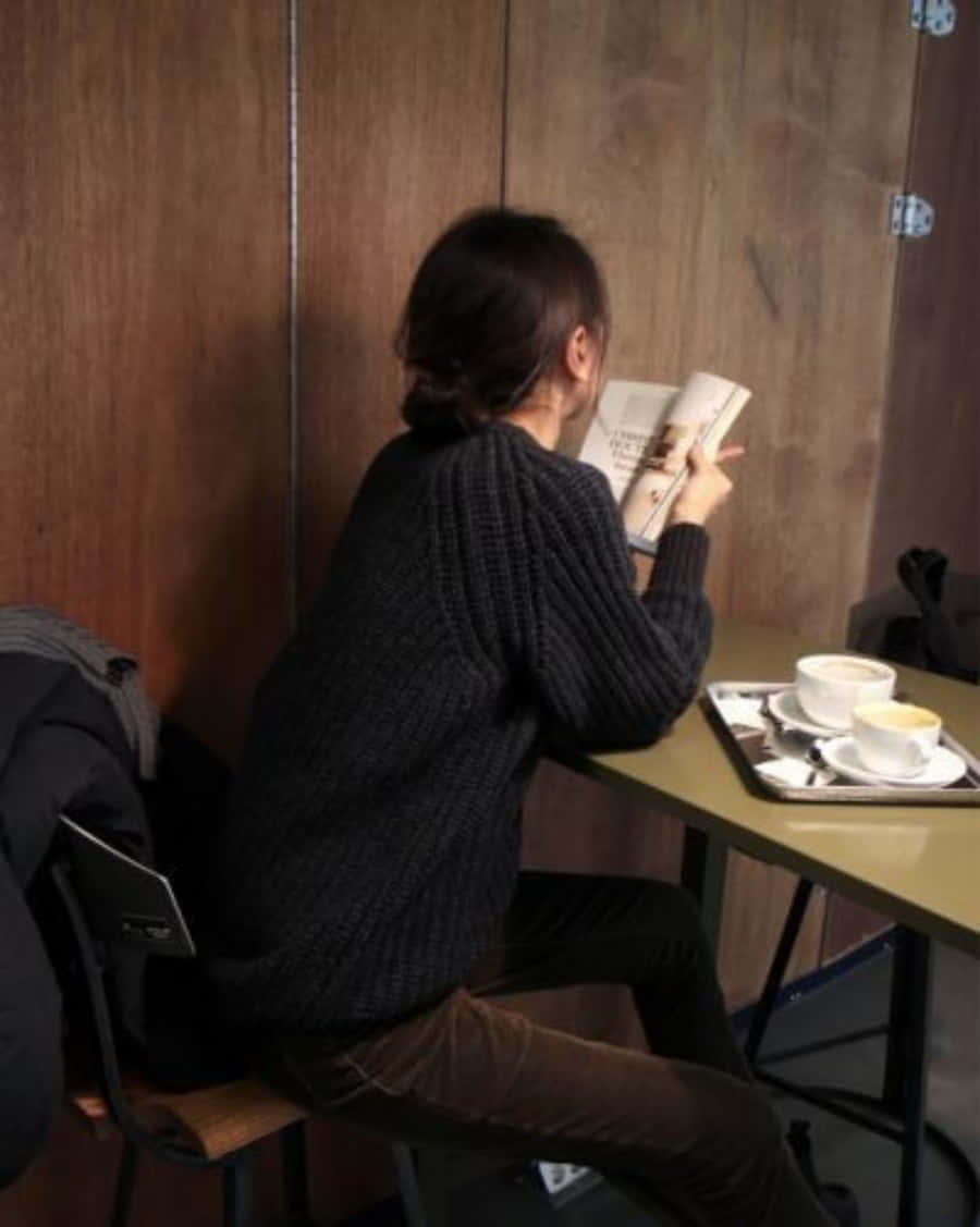 Study Girl 桌布 壁紙 — 一個穿著深色針織毛衣的人在一間舒適的咖啡館裡，在木桌上放著兩杯咖啡，正在閱讀一本書。