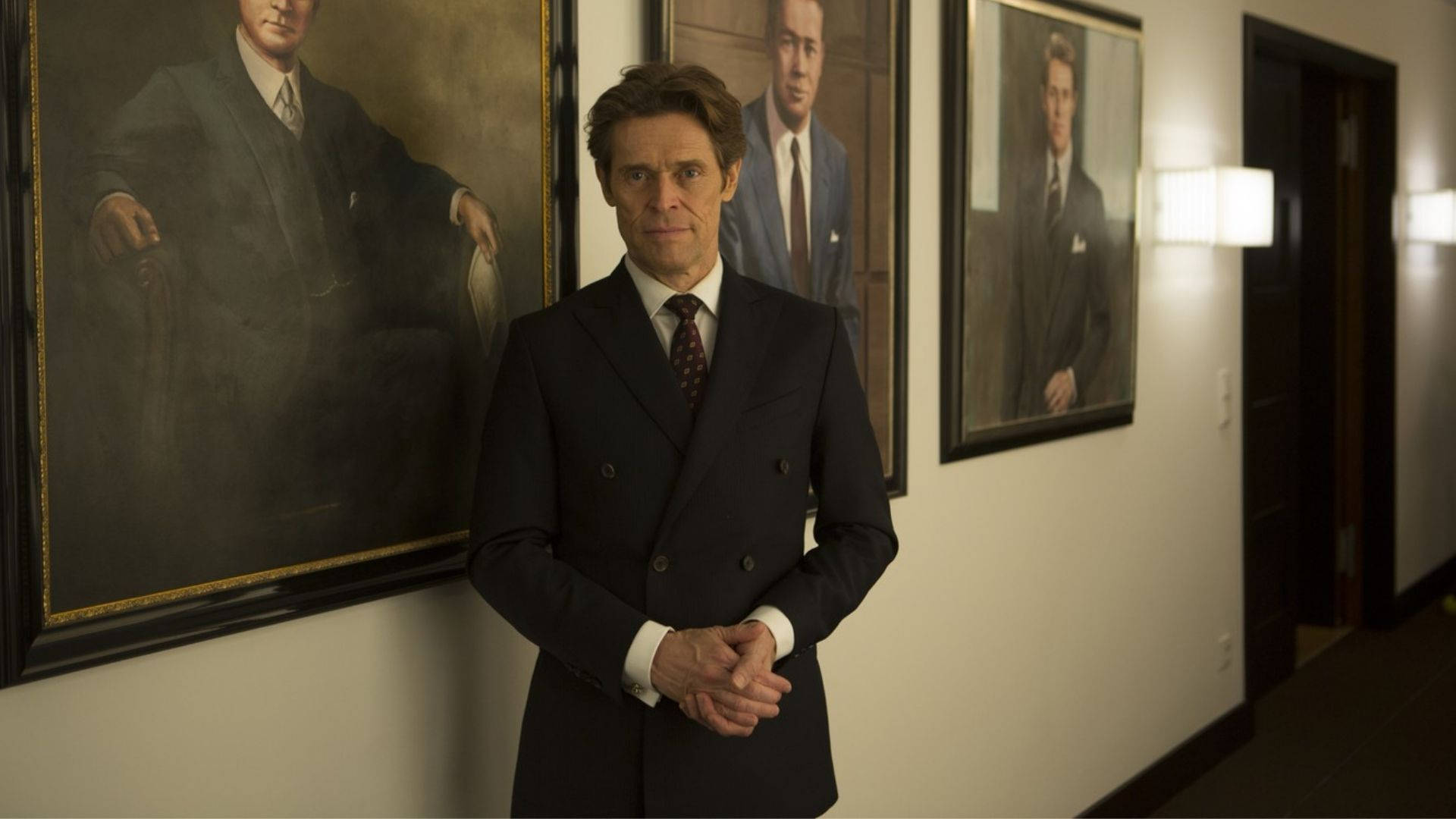 Willem Dafoe 桌布 壁紙 — 一個穿著深色雙排扣西裝的男人站在走廊裡，走廊兩旁掛著鑲框肖像畫，牆上裝有溫暖的壁燈。