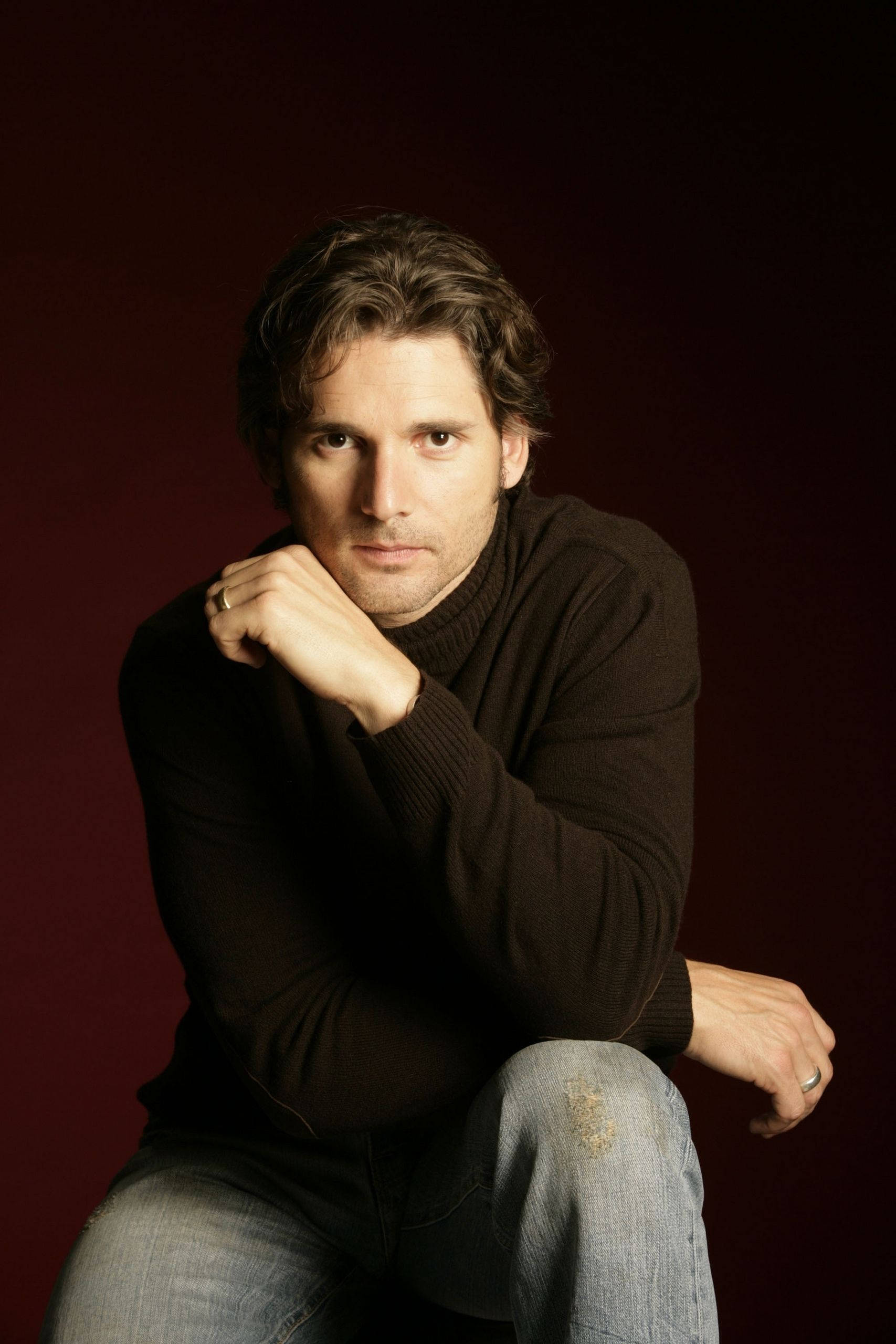 actor 桌布 壁紙 — 一個穿著深色高領毛衣和牛仔褲的男人，在深紅色背景前若有所思地坐著，手托著下巴。