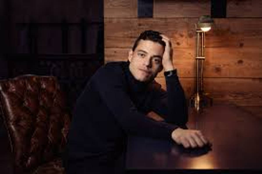 Rami Malek 桌布 壁紙 — 一個穿著深色高領毛衣的男人在木桌上用手托著頭，房間溫暖燈光，有皮椅和壁燈，風格質樸。