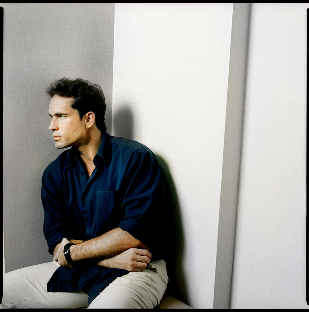 Jason Patric 桌布 壁紙 — 一個穿著深藍色襯衫的男人沉思地坐在純白色的牆前，投下清晰的陰影。