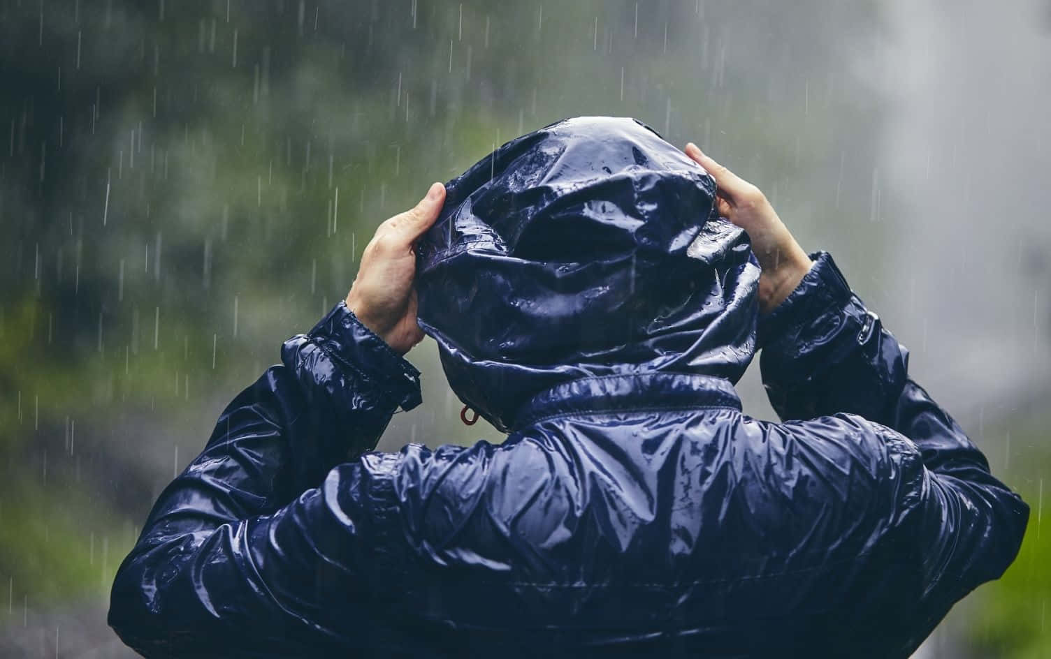 下雨 桌布 壁紙 — 一個穿著深藍色雨衣的人，雙手緊抓布料，將兜帽拉緊抵擋落下的雨水。