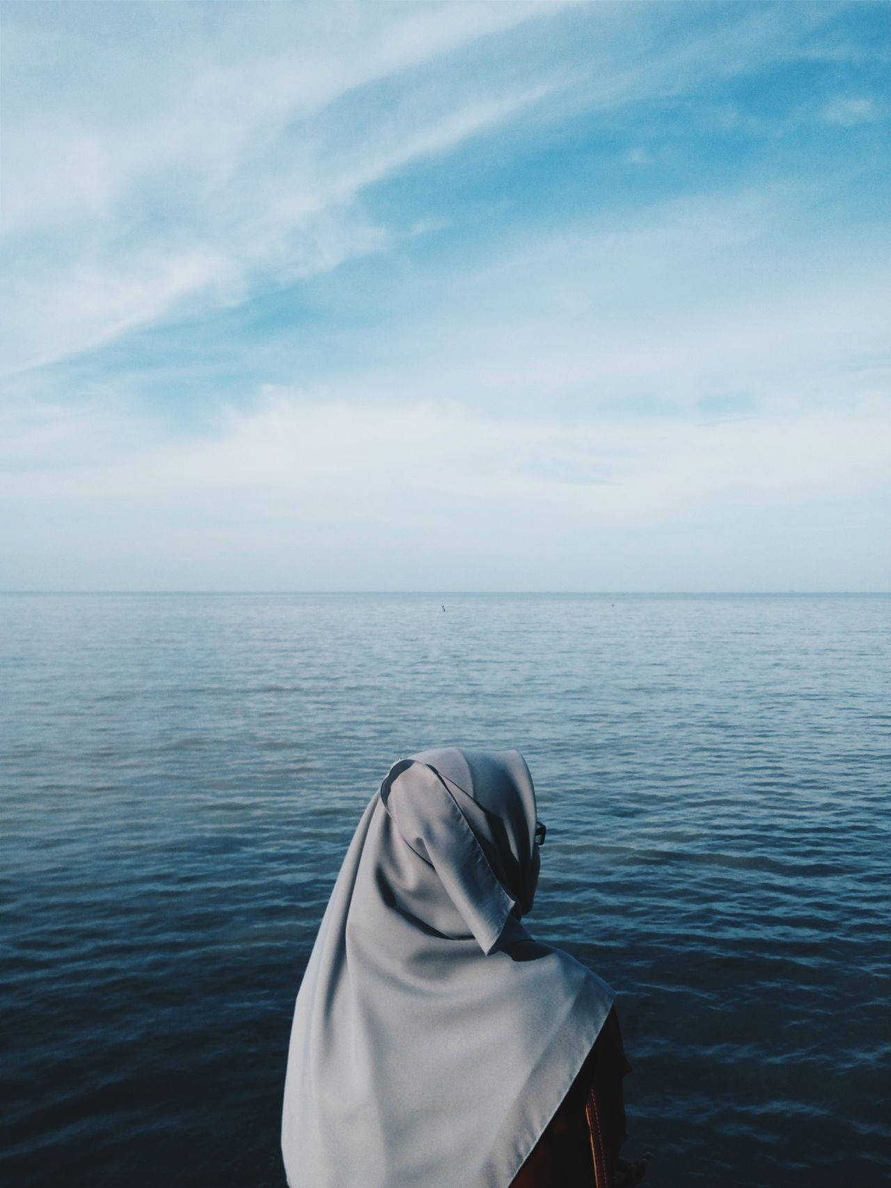 muslim 桌布 壁紙 — 一個穿著淺灰色頭巾的人靜靜地坐著，面對著廣闊平靜的海洋，天空柔和藍色，有縷縷白雲。