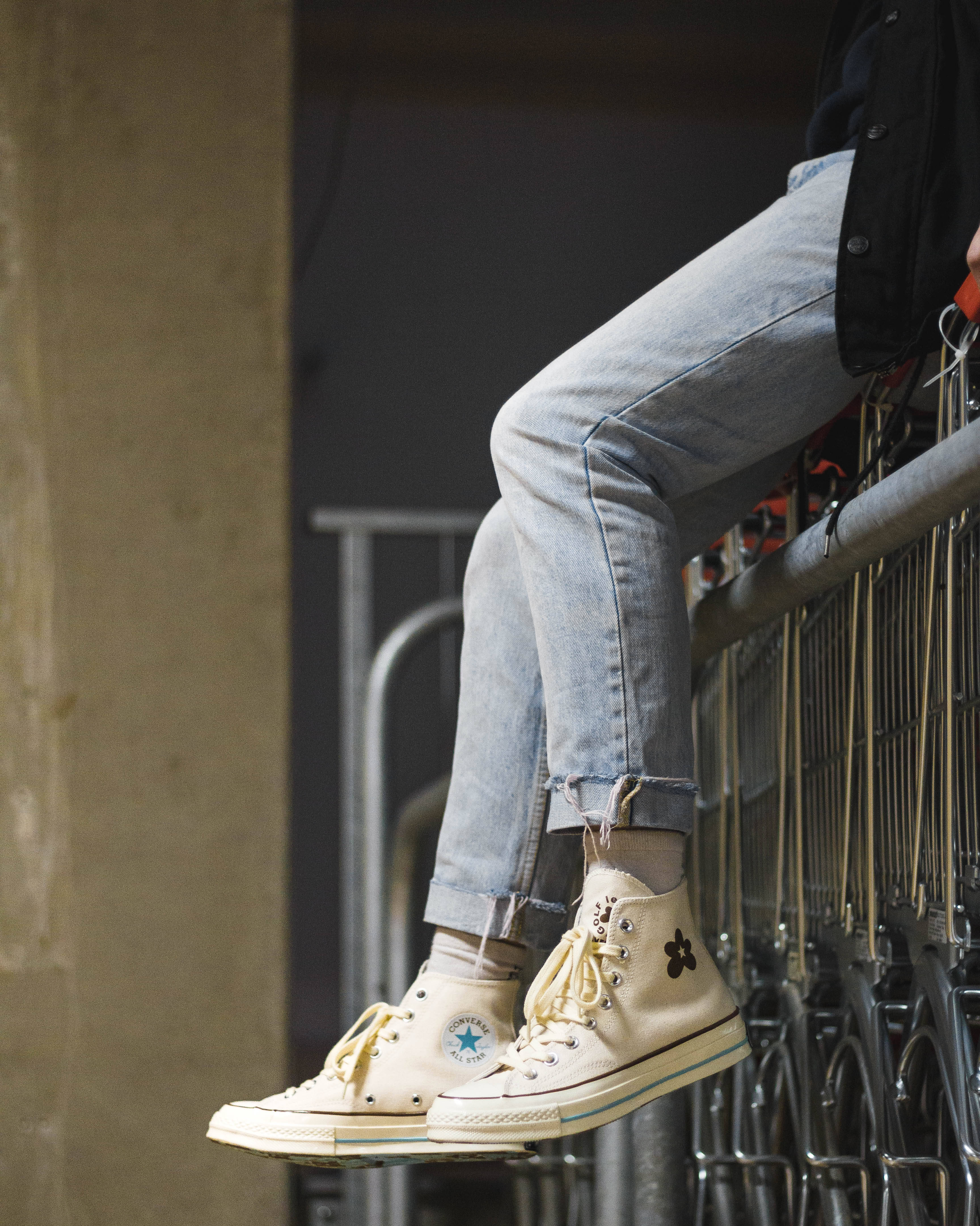 Converse 桌布 壁紙 — 一個穿著淺色牛仔褲和奶油色高筒運動鞋的人，隨意地坐在金屬欄杆上，附近有購物車。