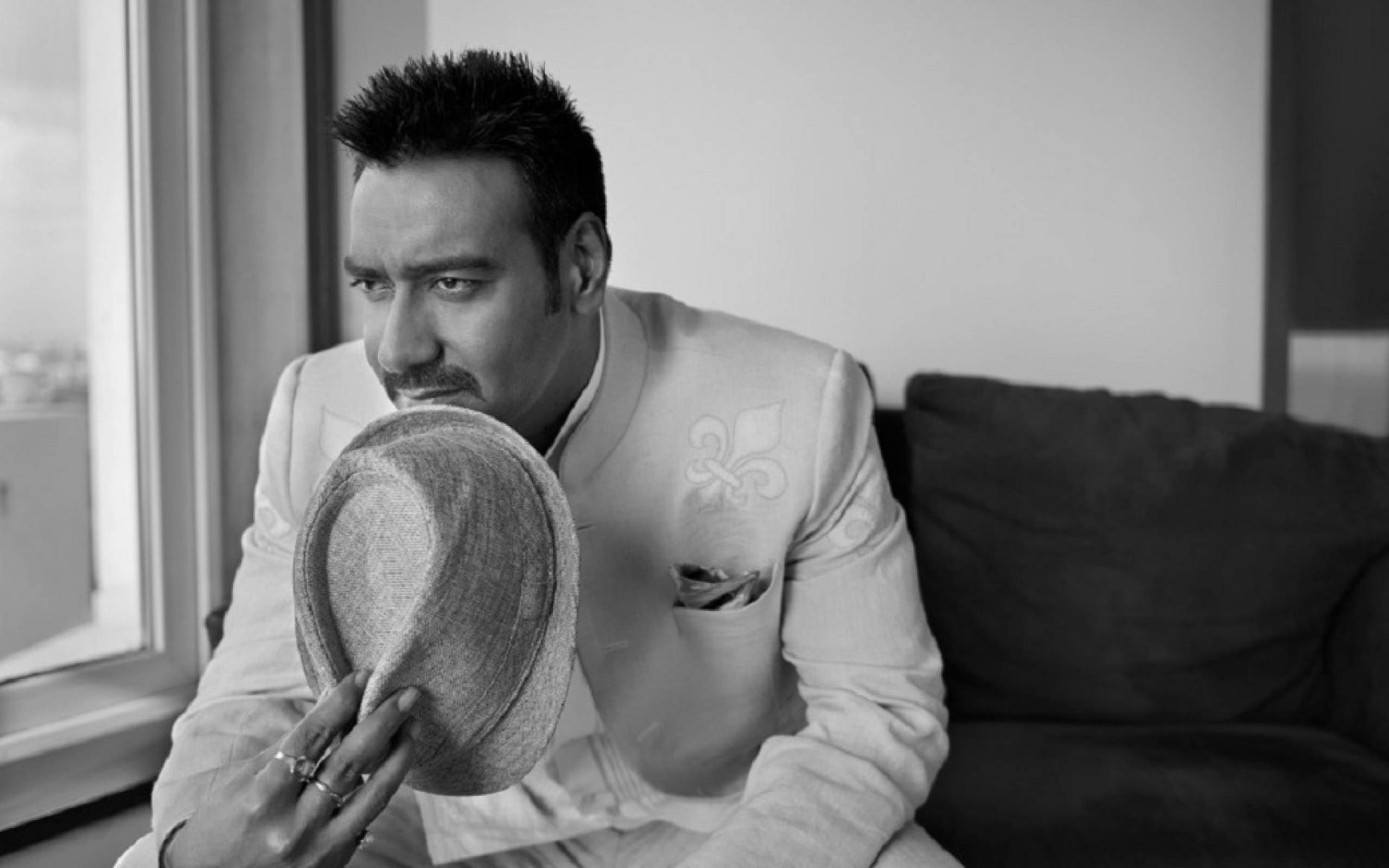 ajay devgn 桌布 壁紙 — 一個穿著淺色西裝的男人在窗邊坐著，手持一頂軟呢帽靠近臉部，黑白肖像照。
