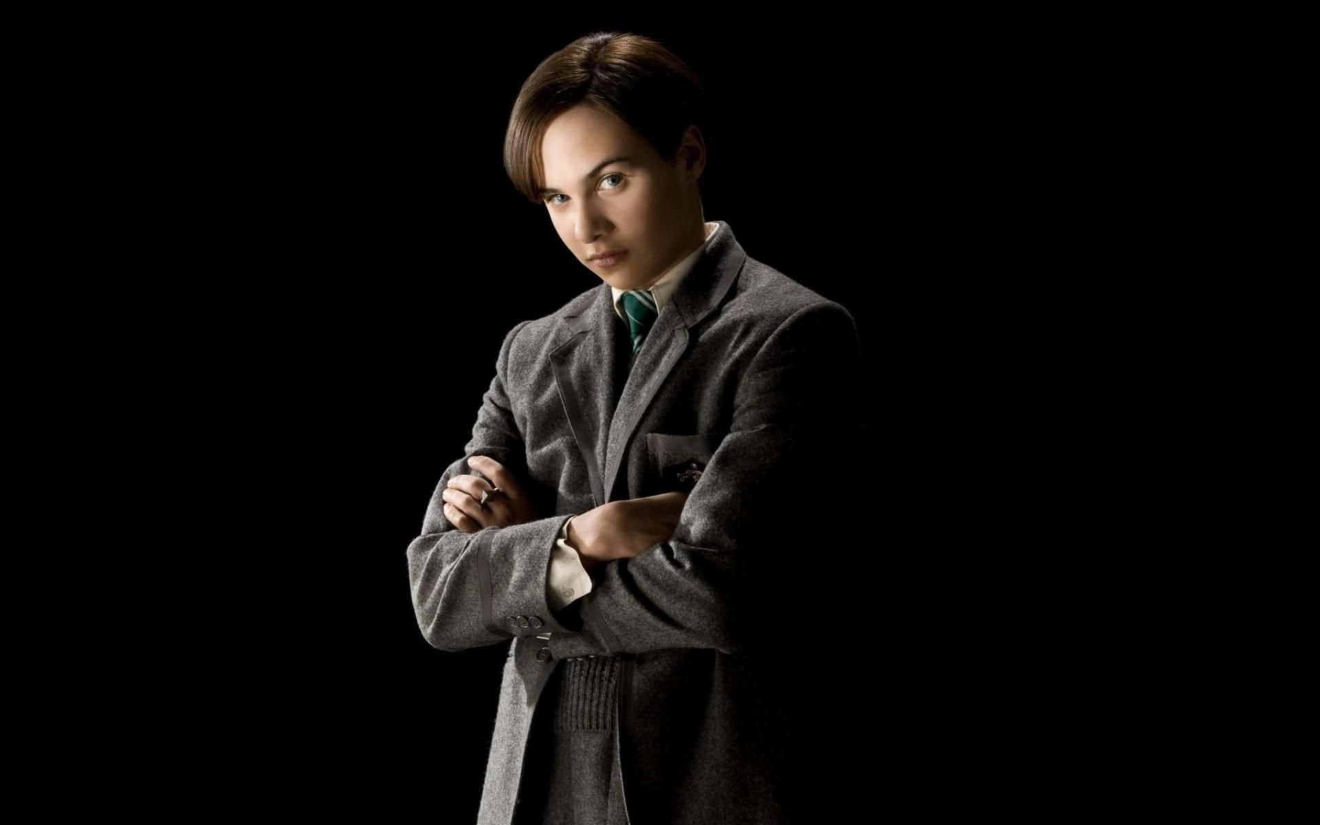 Tom Riddle 桌布 壁紙 — 一個穿著灰色外套的人，雙臂交叉，表情嚴肅，站在黑色背景前。