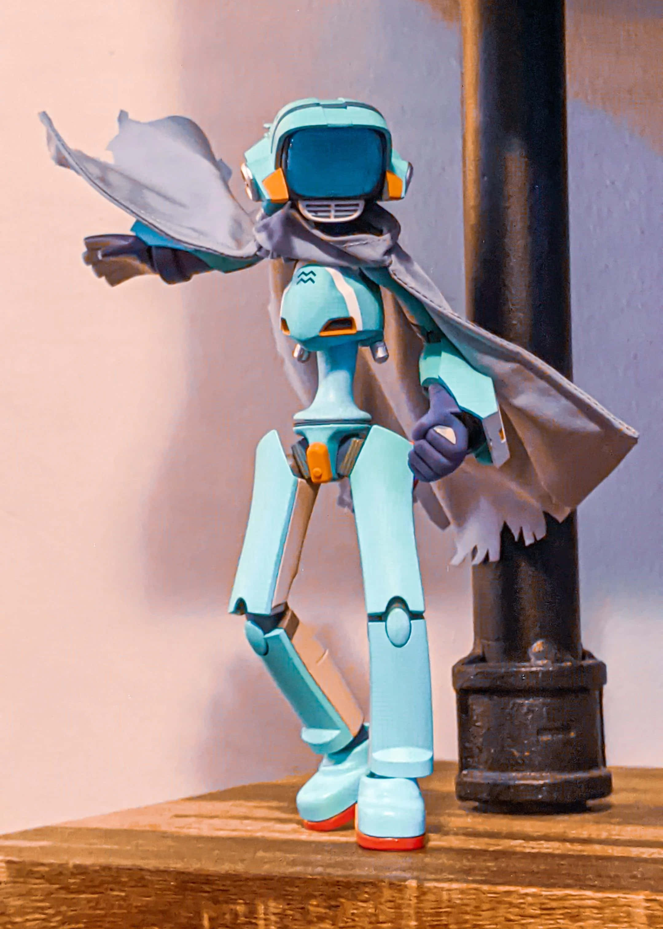 Flcl Canti 桌布 壁紙 — 一個穿著灰色斗篷的藍綠色機器人公仔，英雄式地站在木質表面上，旁邊有一根黑色柱子。