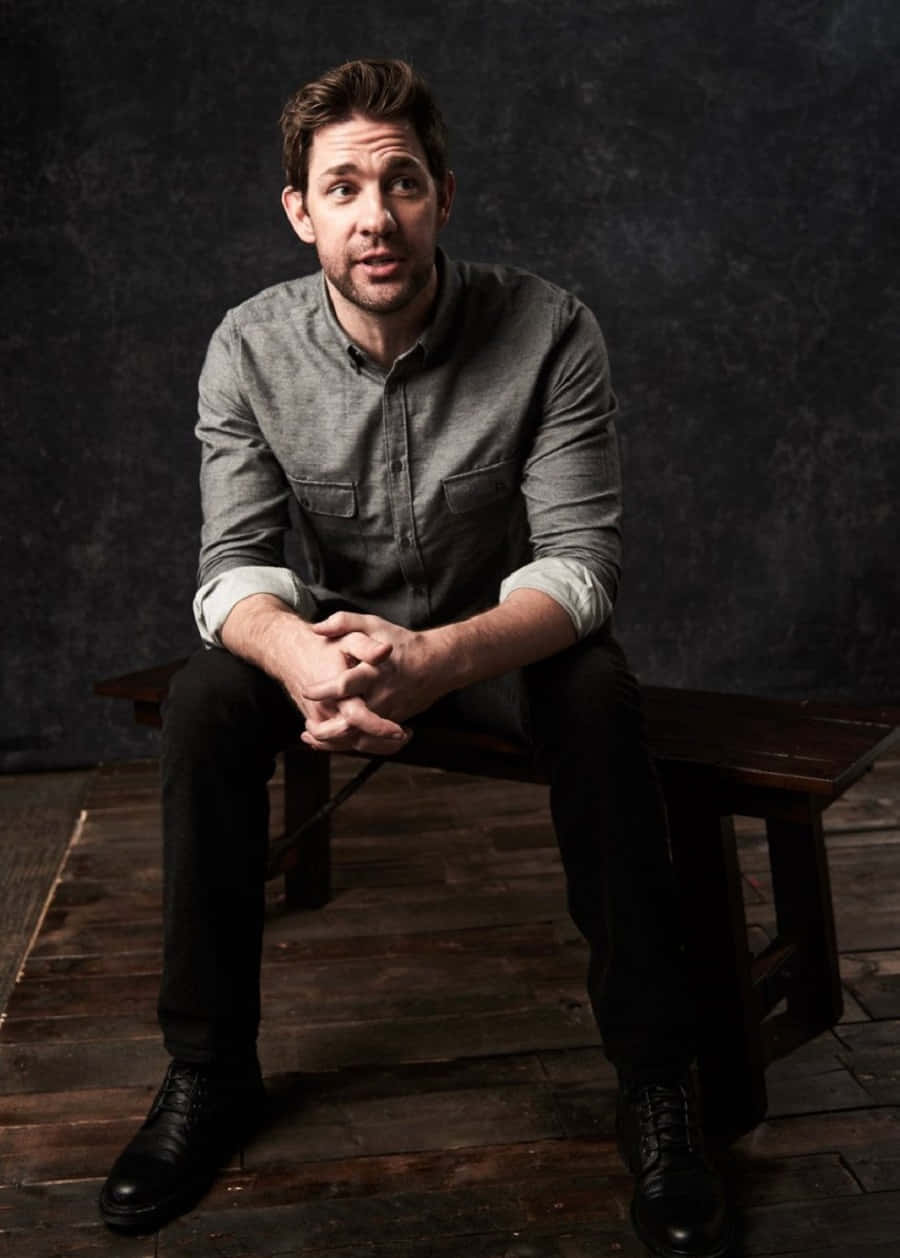 john krasinski 桌布 壁紙 — 一個穿著灰色襯衫的男人坐在木長椅上，靠著深色紋理牆壁沉思。