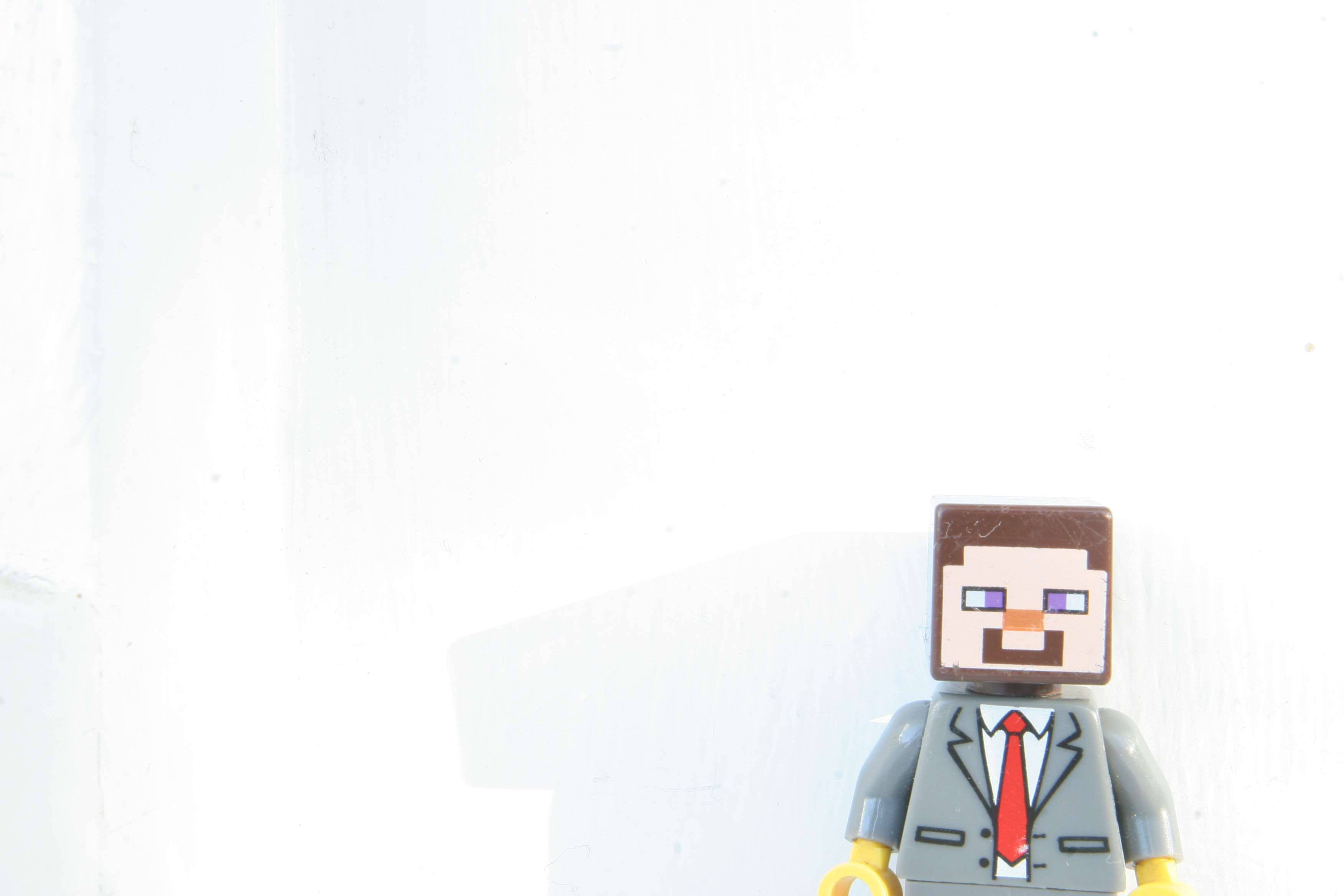 我的世界 史蒂夫 桌布 壁紙 — 一個穿著灰色西裝和紅色領帶的Minecraft風格LEGO迷你人偶站在純白色牆壁前。