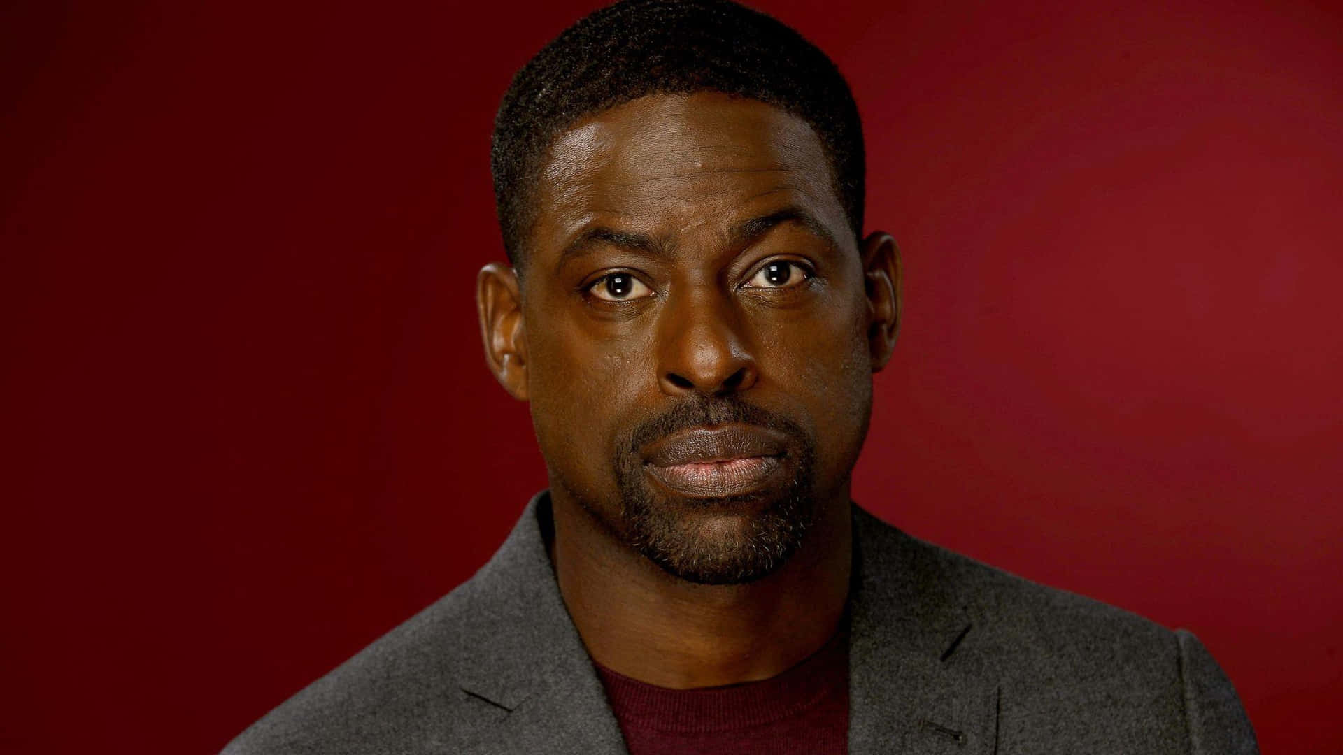Sterling K Brown 桌布 壁紙 — 一個穿著灰色西裝外套和栗色襯衫的男人，在深紅色工作室背景前專注地凝視。