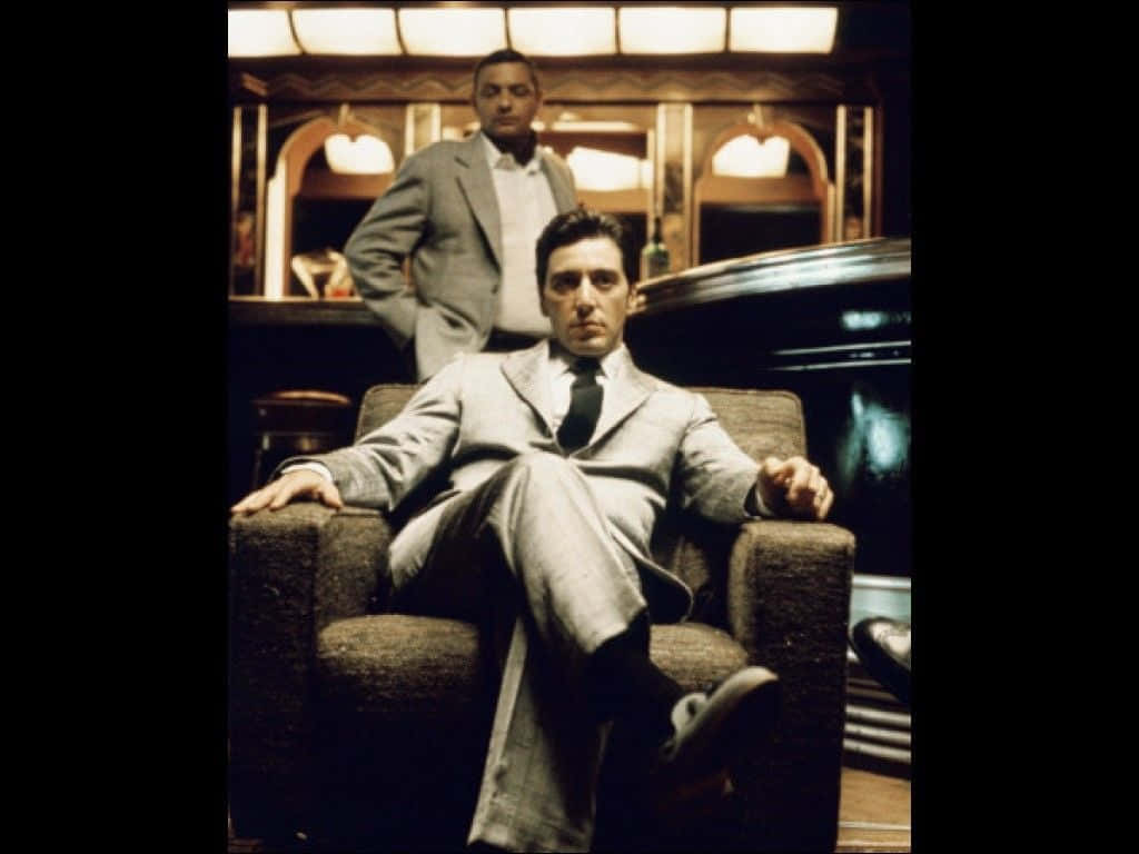 The Godfather 桌布 壁紙 — 一個穿著灰色西裝的男人自信地坐在燈光昏暗的酒吧的椅子上，另一個男人站在他身後。