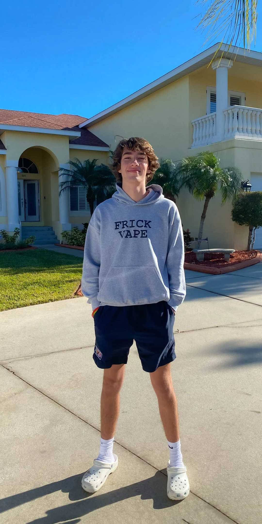 Baylen Levine 桌布 壁紙 — 一個穿著灰色連帽衫和Crocs的年輕男子站在陽光普照的人行道上，對著一棟有棕櫚樹的黃色房子微笑。