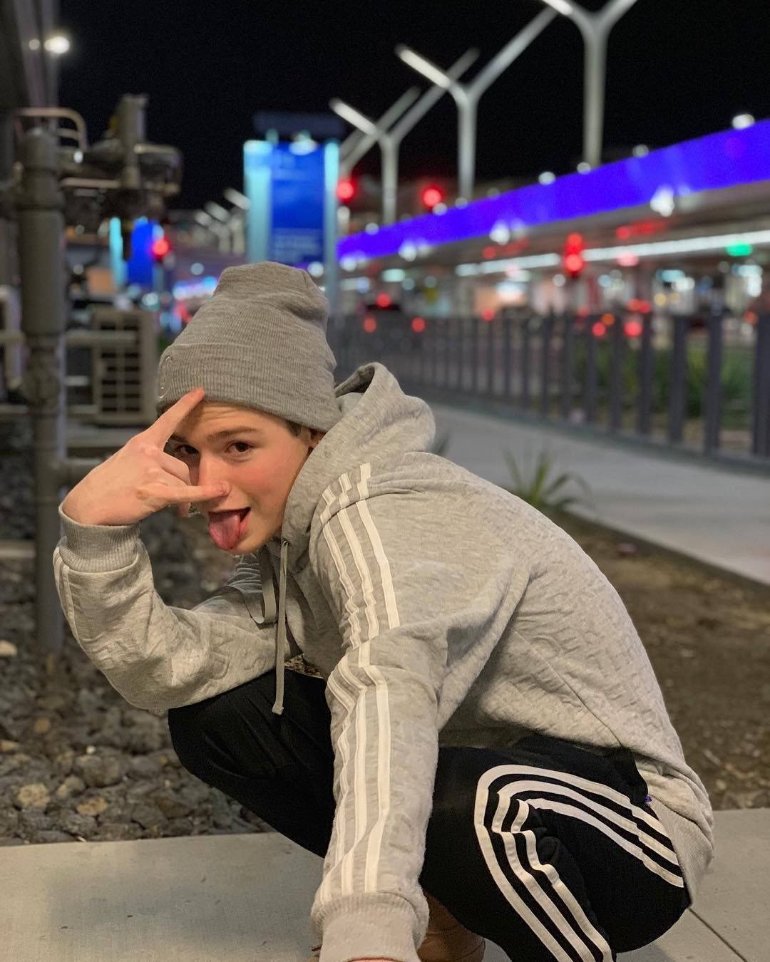 Payton 桌布 壁紙 — 一個穿著灰色連帽衫和毛帽的年輕人，在夜晚頑皮地蹲下，伸出舌頭並做出手勢，背景是燈火通明的城市。