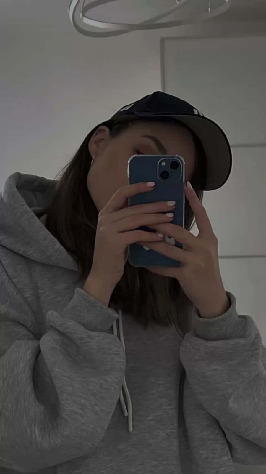 美學 iPhone 壁紙 桌布 壁紙 — 一個穿著灰色連帽衫和黑色帽子的人，在昏暗的室內燈光下用藍色手機拍鏡子自拍。