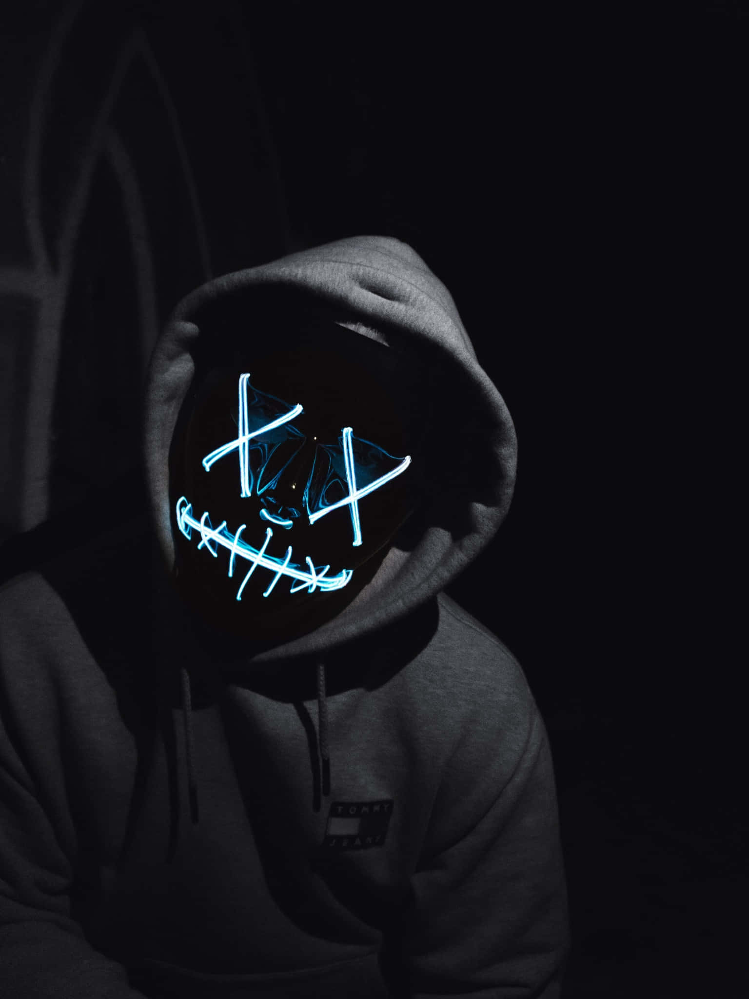 LED 桌布 壁紙 — 一個穿著灰色連帽衫的人，在黑暗背景下戴著發光藍色LED面具，帶有縫合微笑和X眼睛。