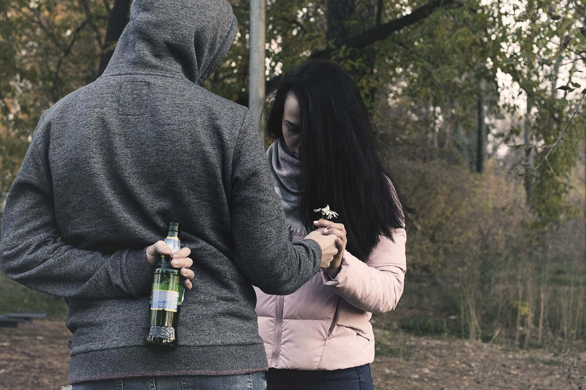 Sad Couple 桌布 壁紙 — 一個穿著灰色連帽衫的人拿著啤酒瓶，同時擁抱一個穿著粉紅色外套、緊握著一朵小白花的女人，在一個寧靜的公園裡。