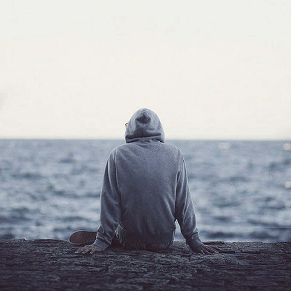 Alone 桌布 壁紙 — 一個穿著灰色連帽衫的人獨自坐在岩石懸崖上，俯瞰著陰天下一片廣闊平靜的海洋。