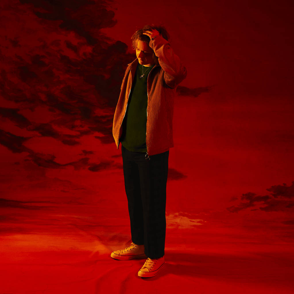Lewis Capaldi 桌布 壁紙 — 一個穿著燈芯絨外套的男人獨自站在超現實的紅色天空下，烏雲密布，手放在頭上沉思。