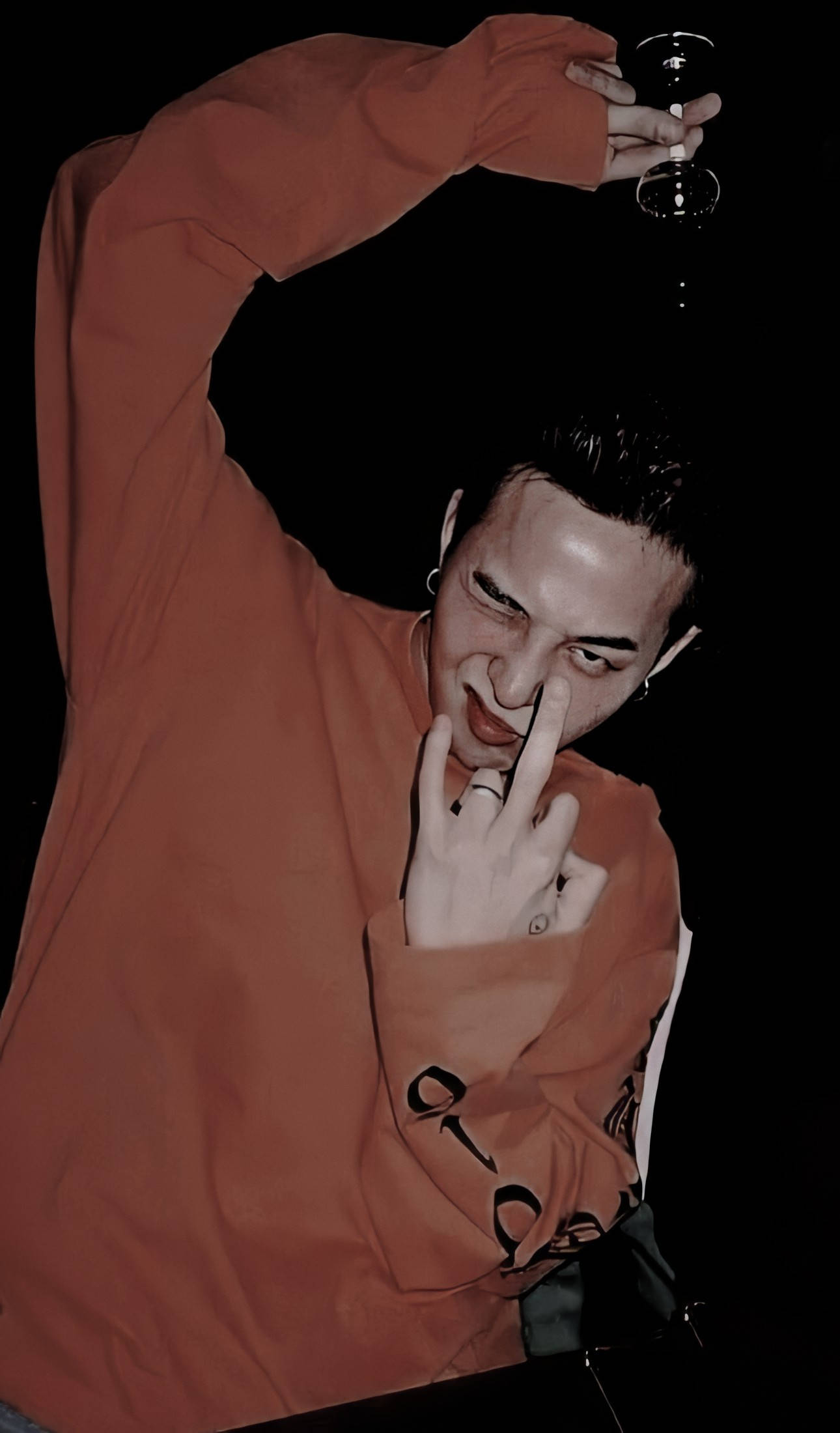 G-dragon 桌布 壁紙 — 一個穿著燒焦橙色連帽衫的男人拿著一杯酒，在黑色背景前頑皮地觸摸鼻子。