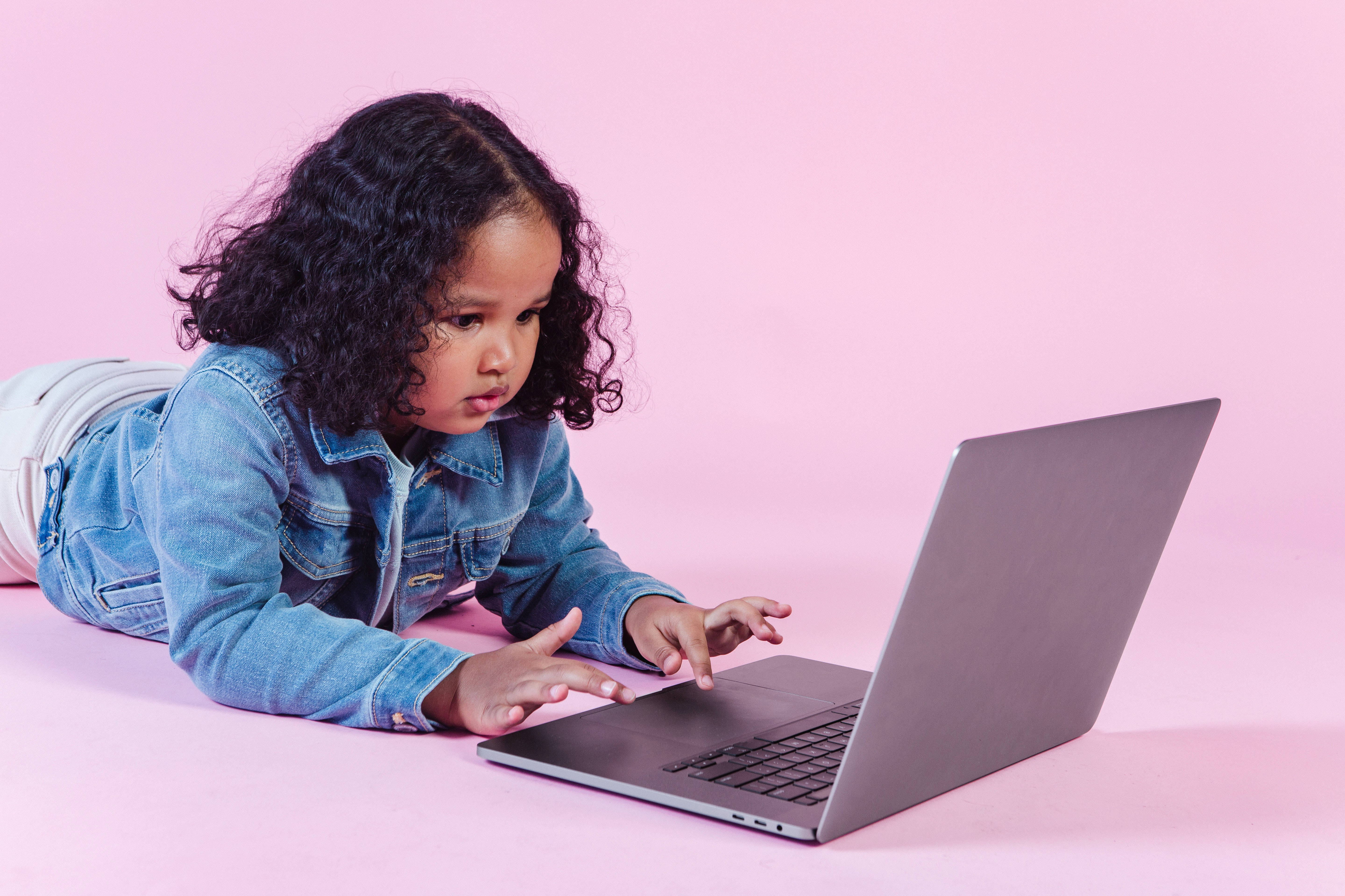 Cute Laptop 桌布 壁紙 — 一個穿著牛仔外套、捲髮的小女孩躺在粉紅色地板上，專注地使用銀色筆記型電腦。