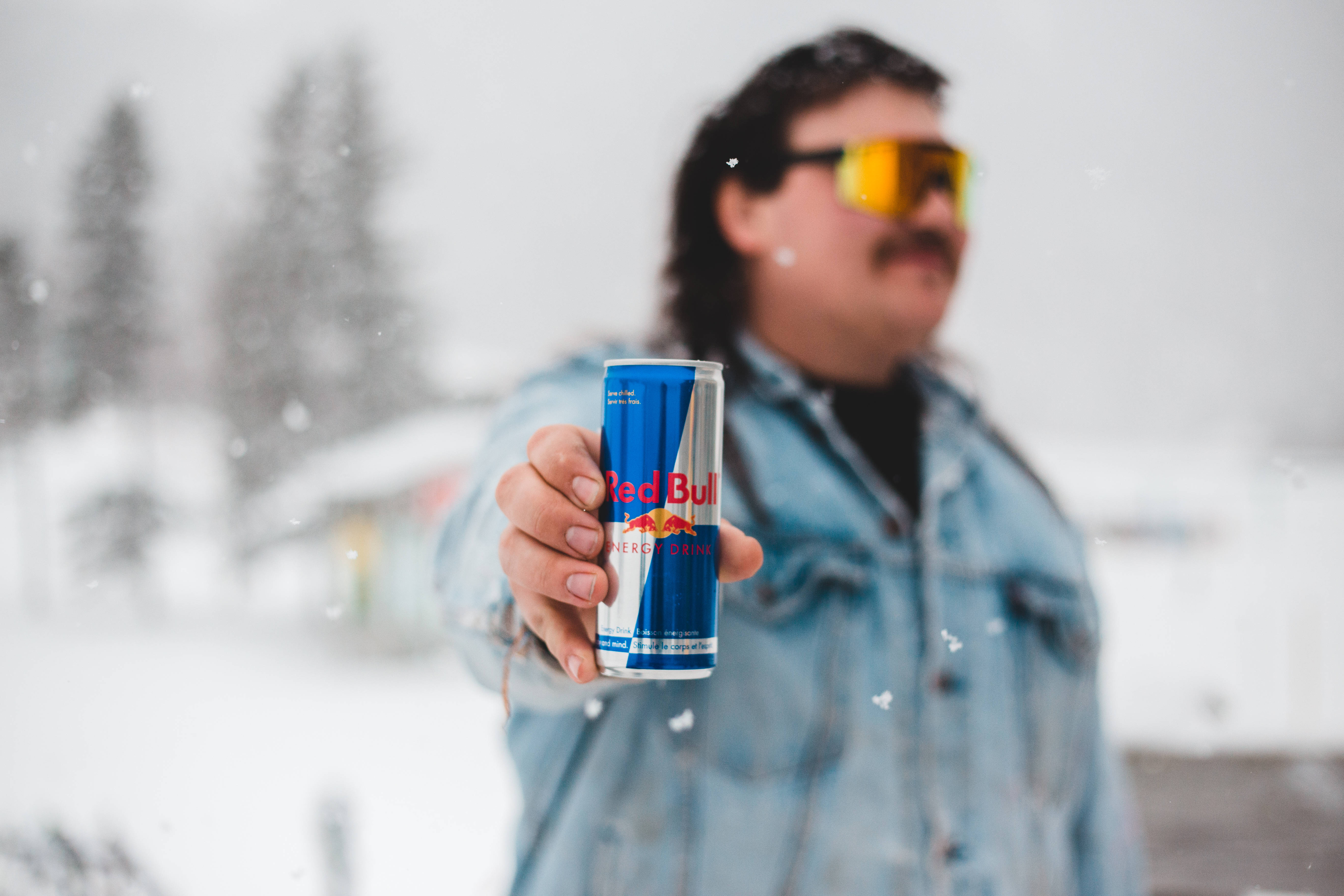 bull 桌布 壁紙 — 一個穿著牛仔外套和黃色護目鏡的男人在雪景中伸出紅牛罐，雪花飄落。