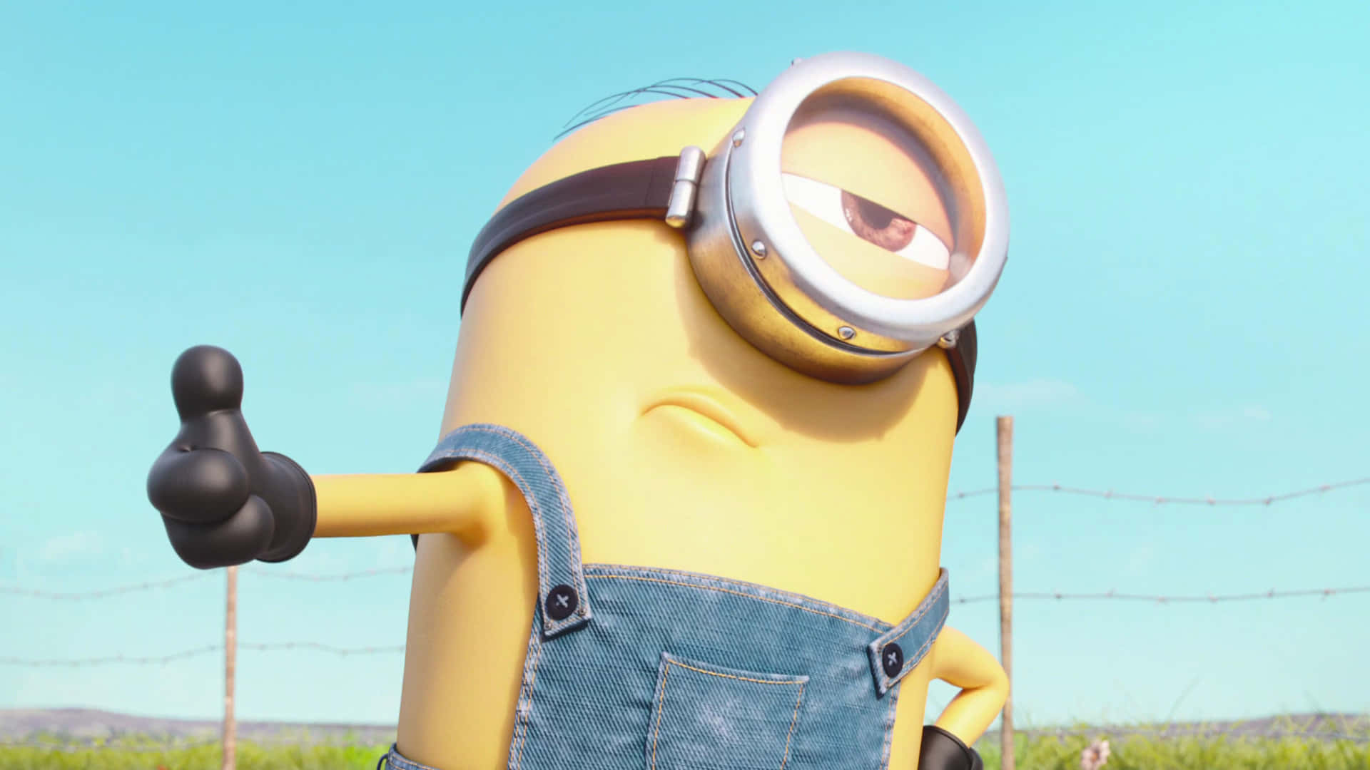 Thumbs Up 桌布 壁紙 — 一個穿著牛仔工裝褲的黃色Minion在明亮的藍天和鐵絲網圍欄前豎起大拇指。