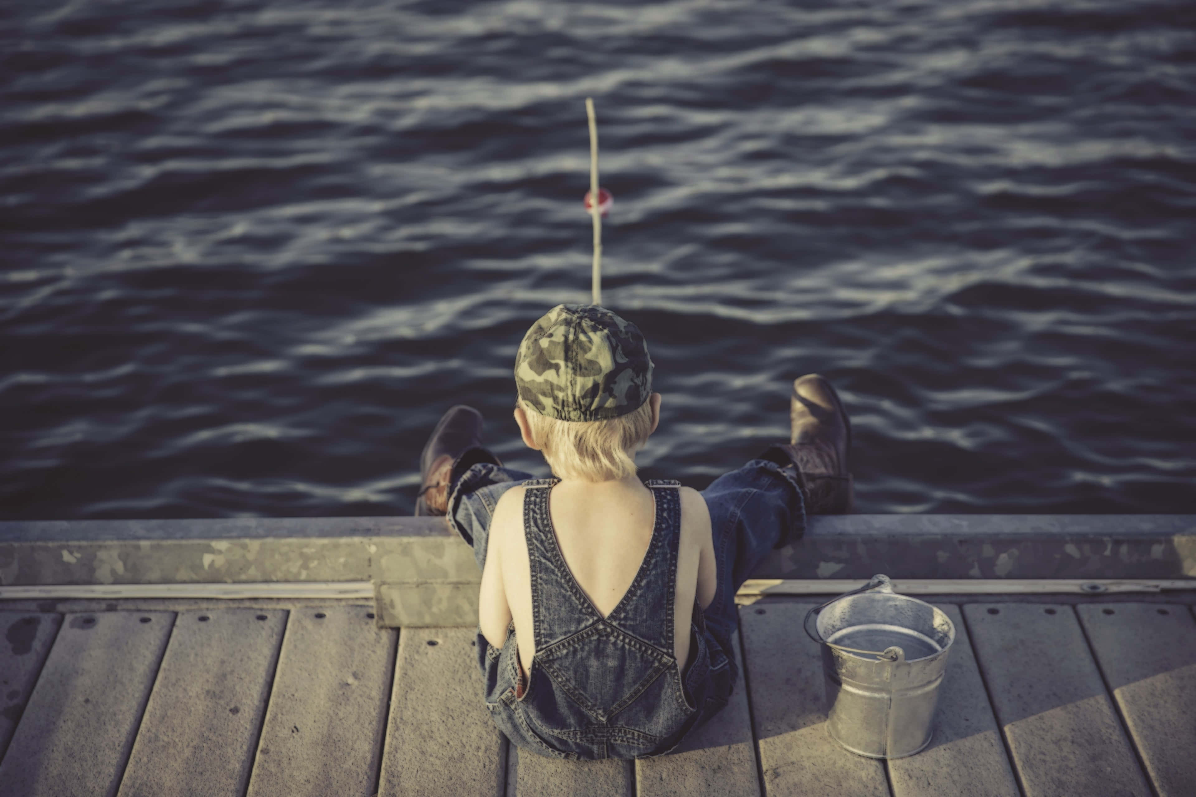 Fishing Bait 桌布 壁紙 — 一個穿著牛仔布工裝褲和迷彩帽的小孩坐在木碼頭上釣魚，浮標在平靜的水面上晃動。