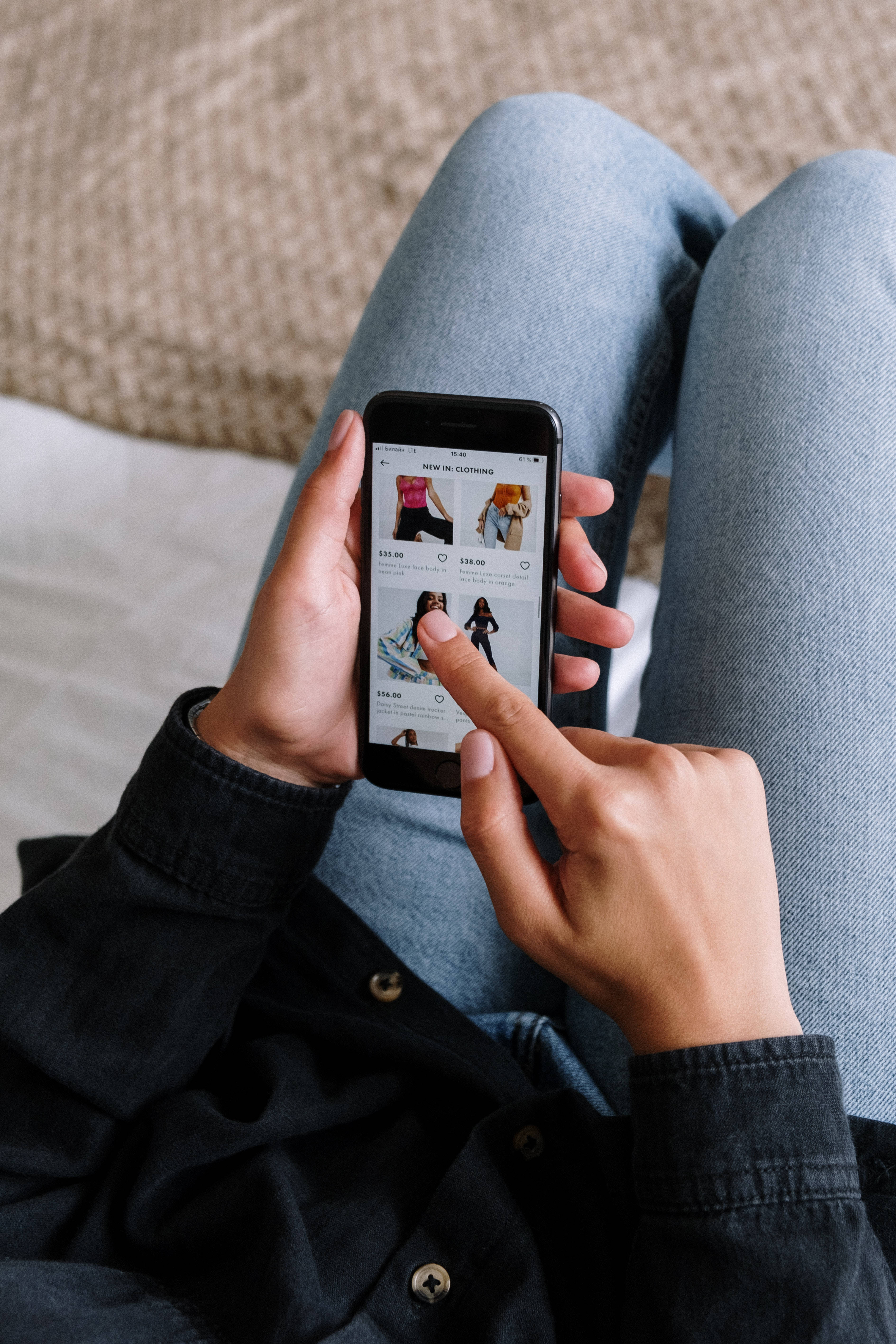 酷炫iPhone 桌布 壁紙 — 一個穿著牛仔褲和深色襯衫的人，在帶有紋理的沙發上放鬆時，瀏覽智慧型手機上的服裝應用程式。