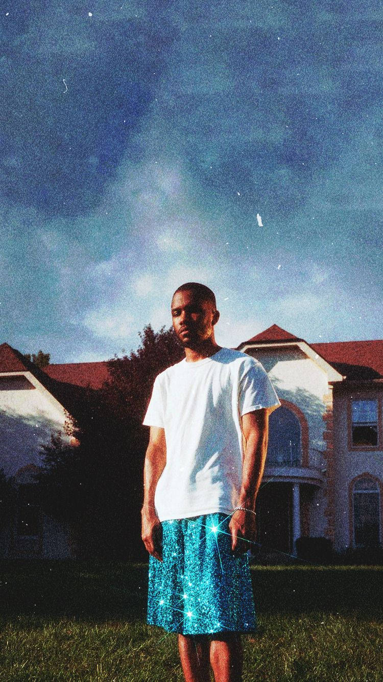 Frank Ocean 桌布 壁紙 — 一個穿著白色T恤和閃亮藍色短褲的男人站在草地庭院中，面前是郊區房屋，背景是戲劇性的黃昏天空。