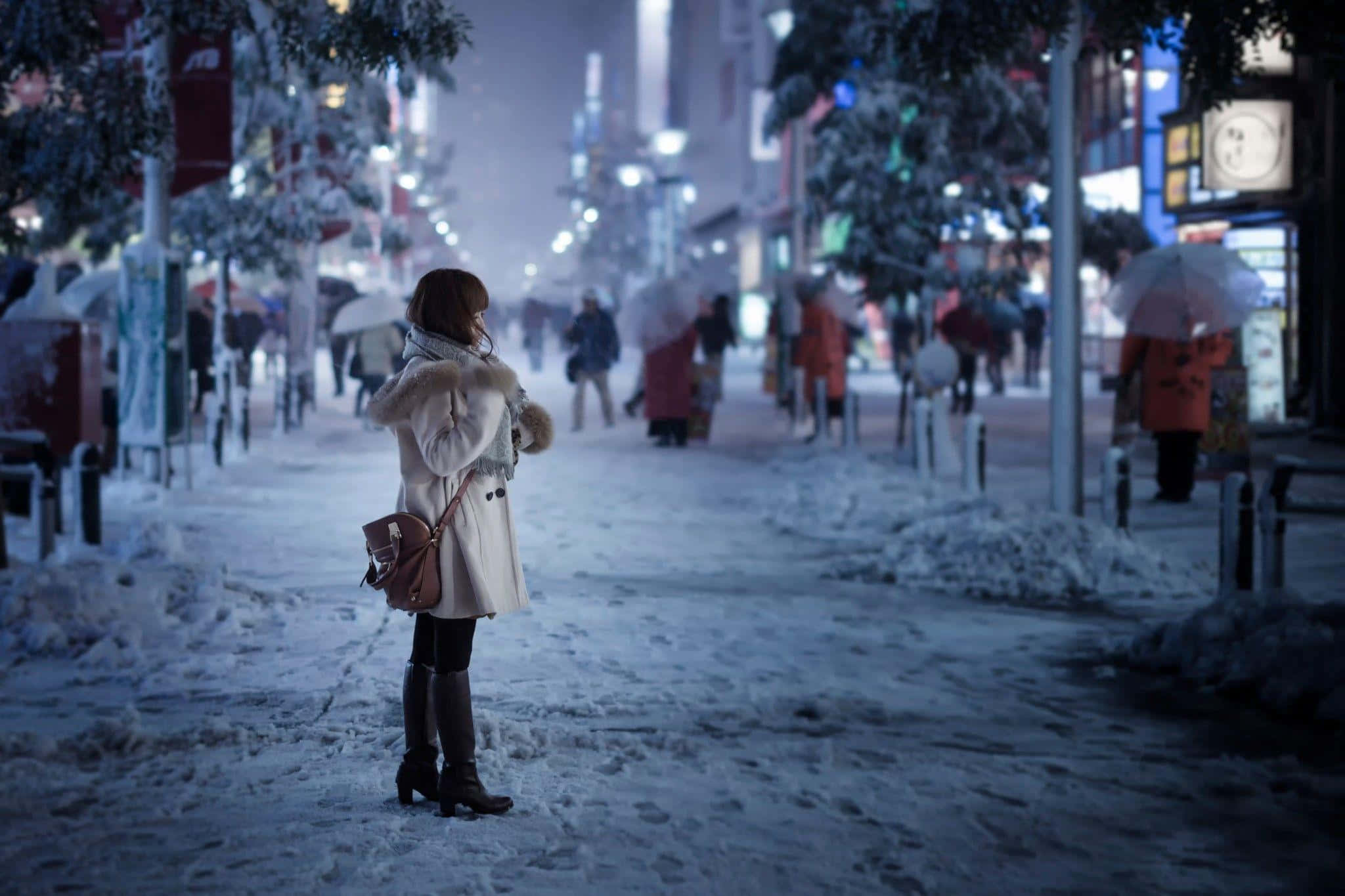 Winter Cityscape 桌布 壁紙 — 一個穿著白色外套的孤獨女人夜晚站在積雪的城市街道上，周圍是模糊的行人和閃爍的燈光。