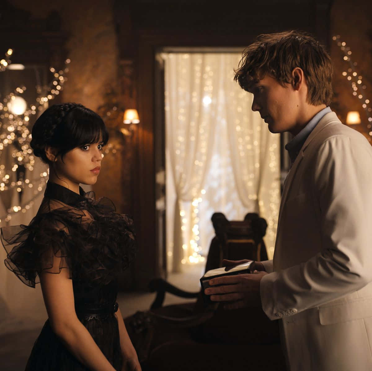 洋裝 桌布 壁紙 — 一個穿著白色外套的男人在昏暗的房間裡，在溫暖的串燈下，向一個穿著黑色連衣裙的女人遞上一個盒子。