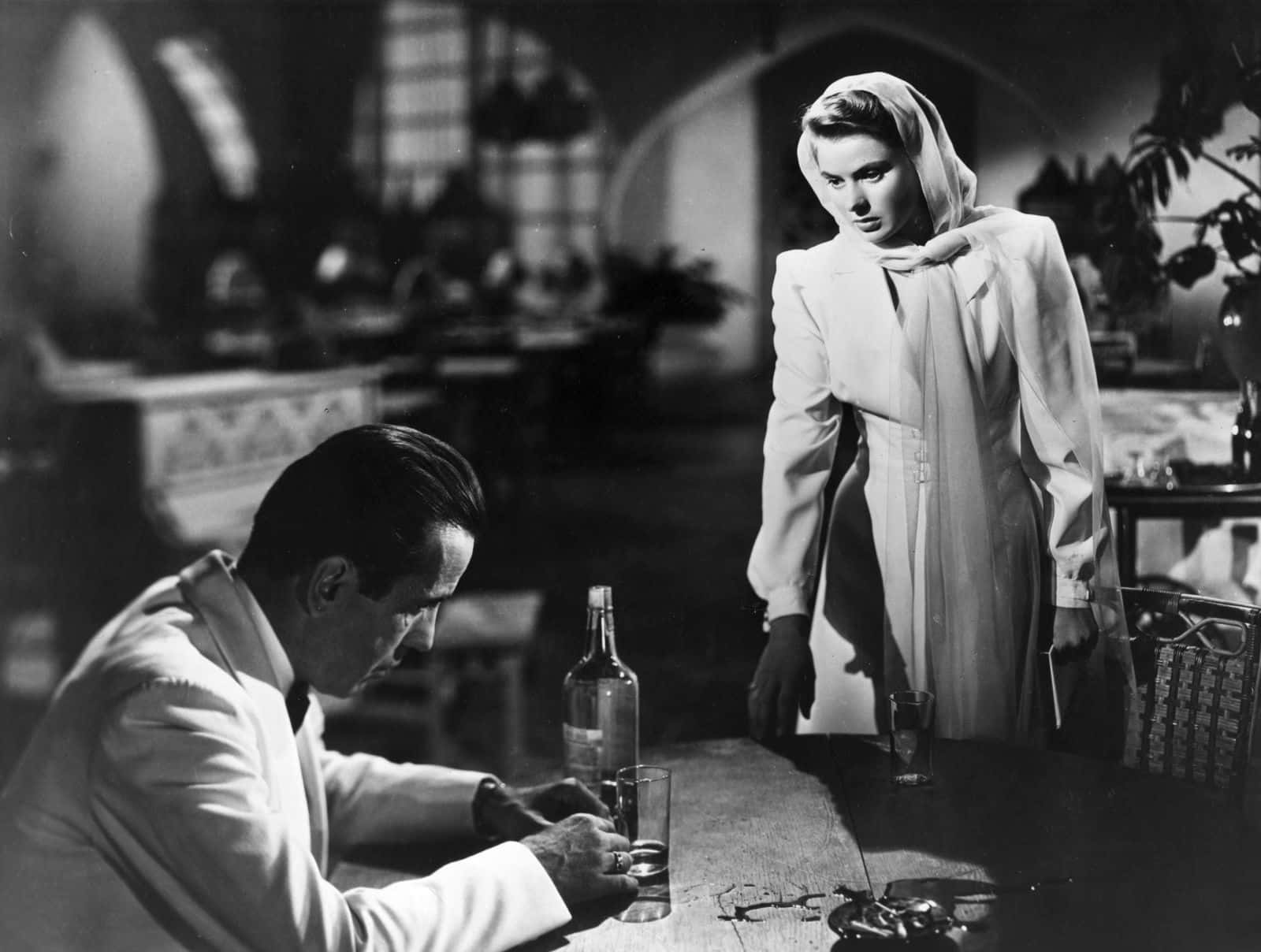 Black And White Movie 桌布 壁紙 — 一個穿著白色外套的男人坐在桌旁倒飲料，而一個穿著白色長袍的女人站在他上方，在一個燈光昏暗的房間裡。