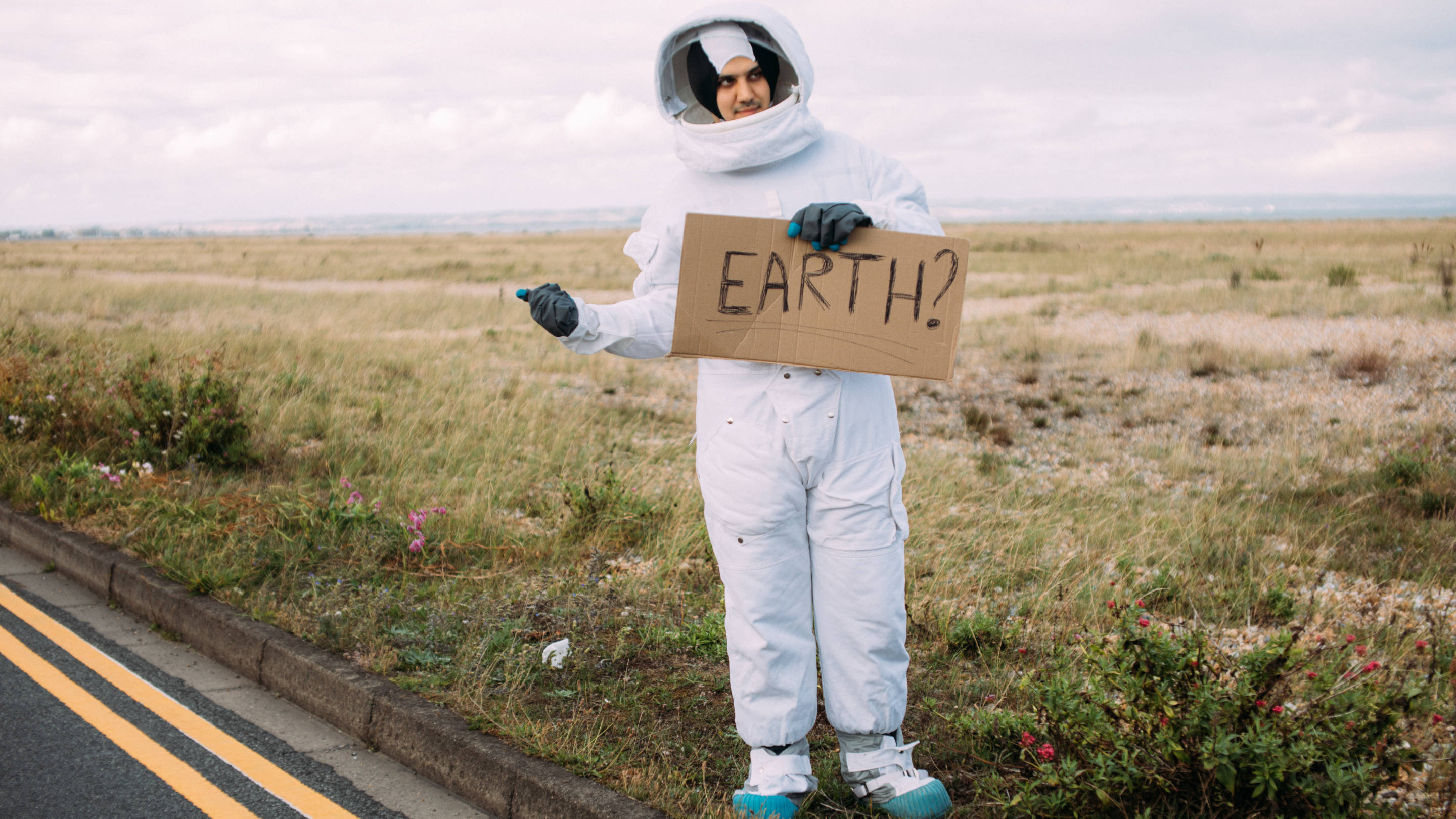 太空人 桌布 壁紙 — 一個穿著白色太空衣的人站在路邊，舉著一個寫著「地球？」的紙板標語。